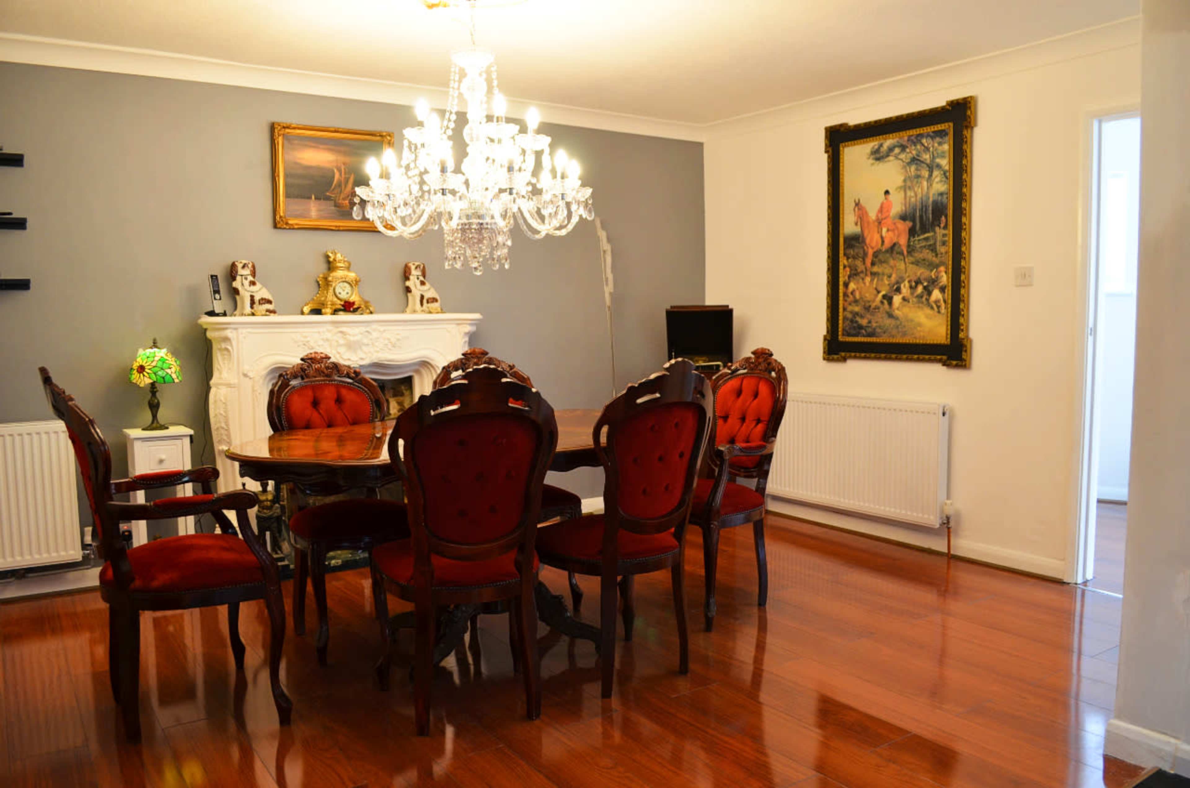 A dining room with a round wooden table surrounded by red upholstered chairs, a chandelier above, and a fireplace with decorative items.