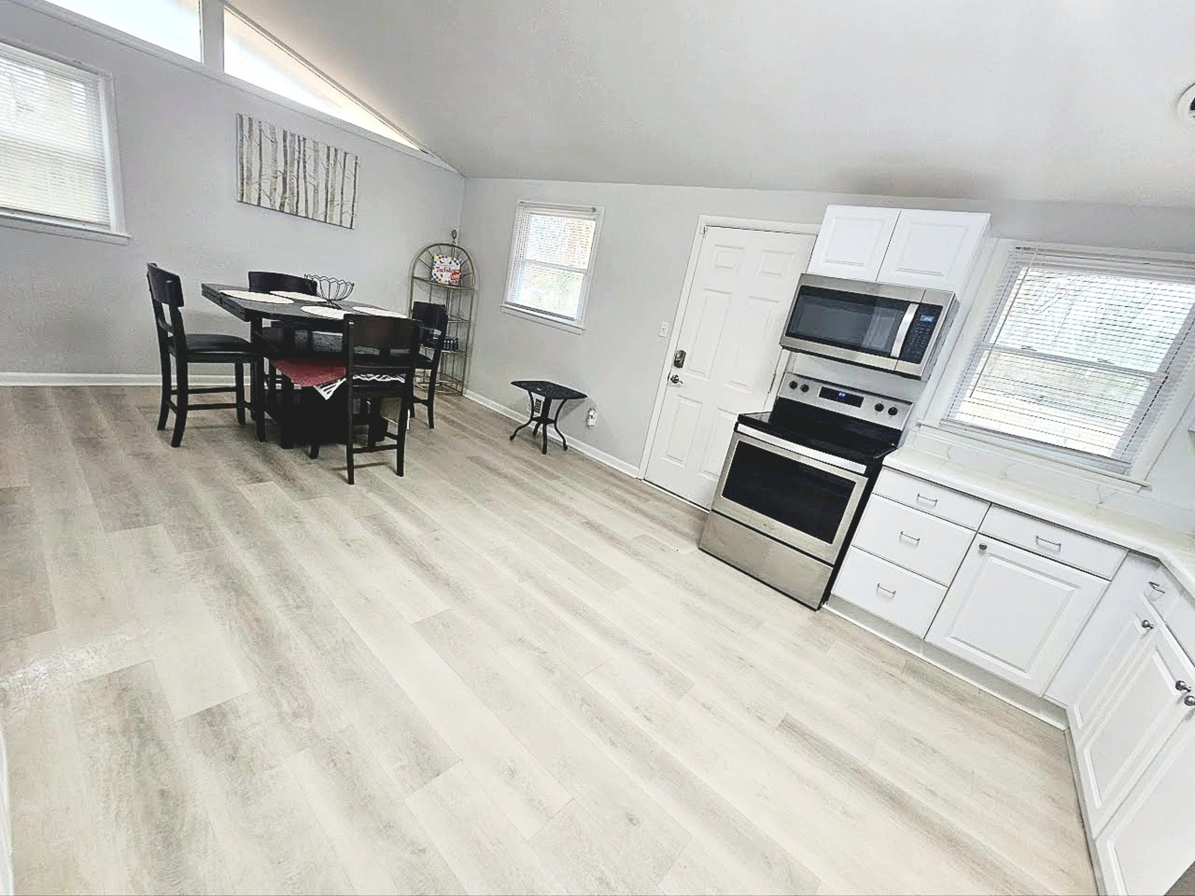 The image shows a bright, modern kitchen and dining area featuring a table with four chairs, stainless steel appliances, and light-colored flooring.