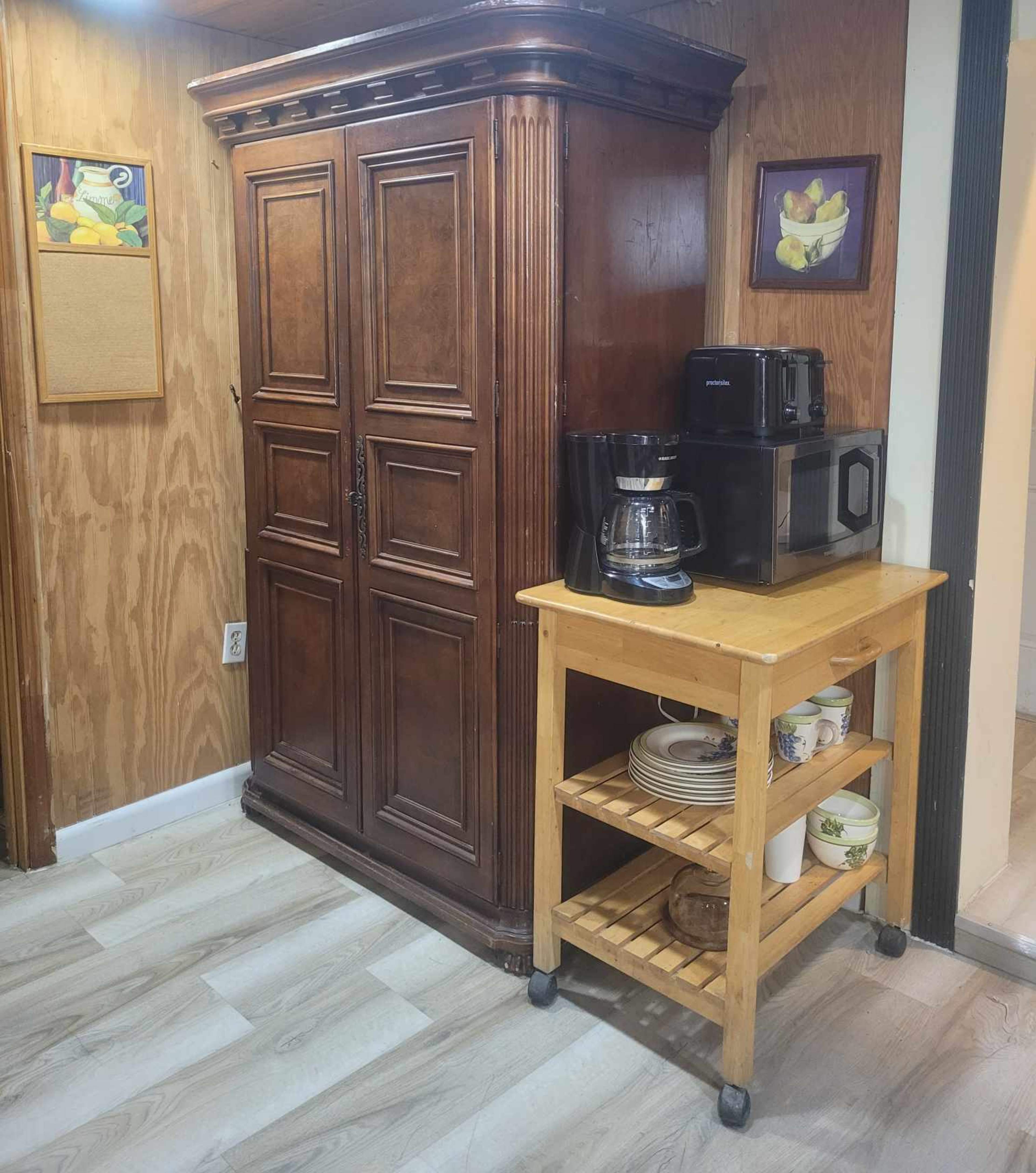 A wooden cabinet stands against a wall next to a rolling cart with dishes and a coffee maker, while a microwave is positioned on top of the cabinet.
