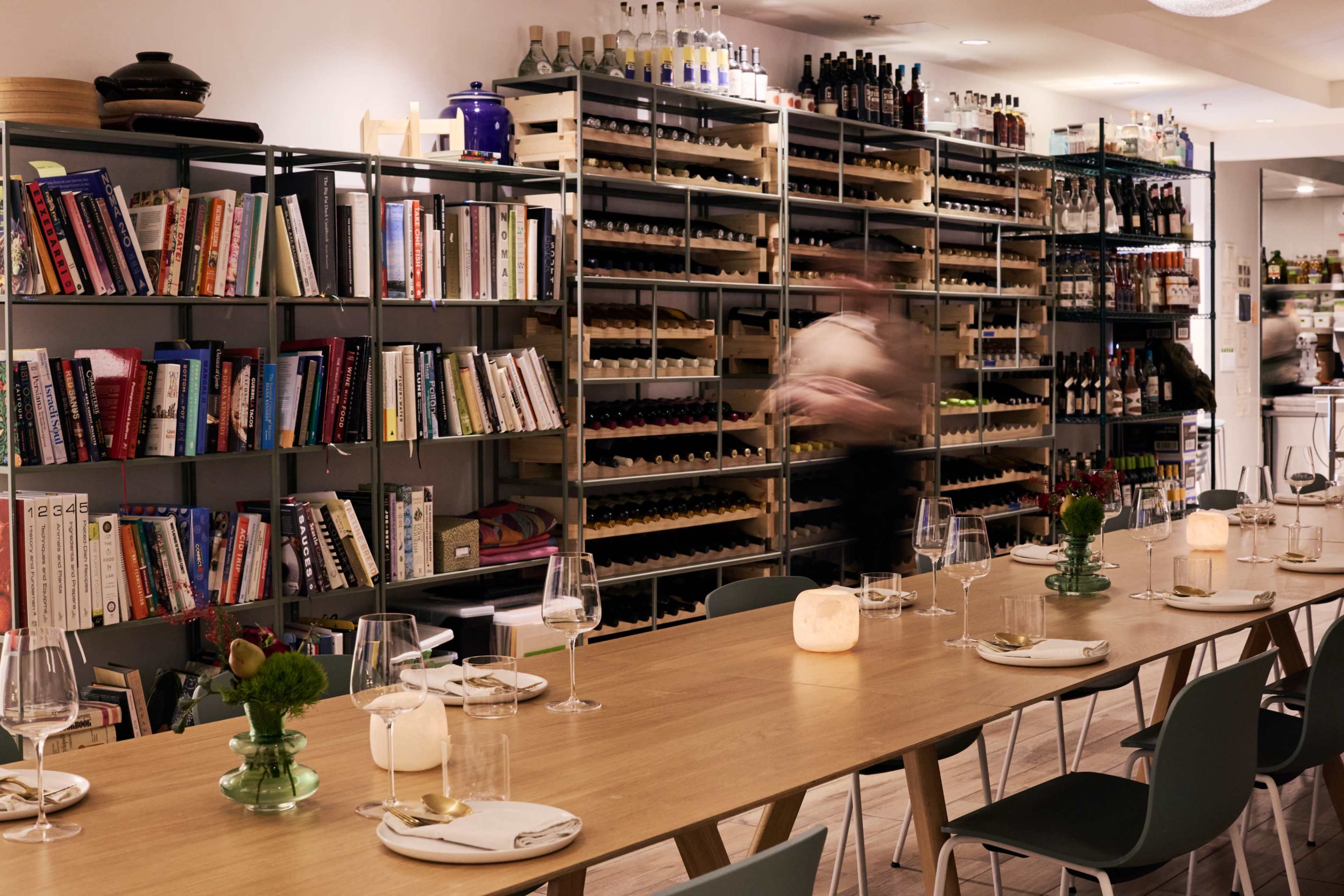 A long wooden table is set for dining in a restaurant with shelves filled with books and bottles in the background.