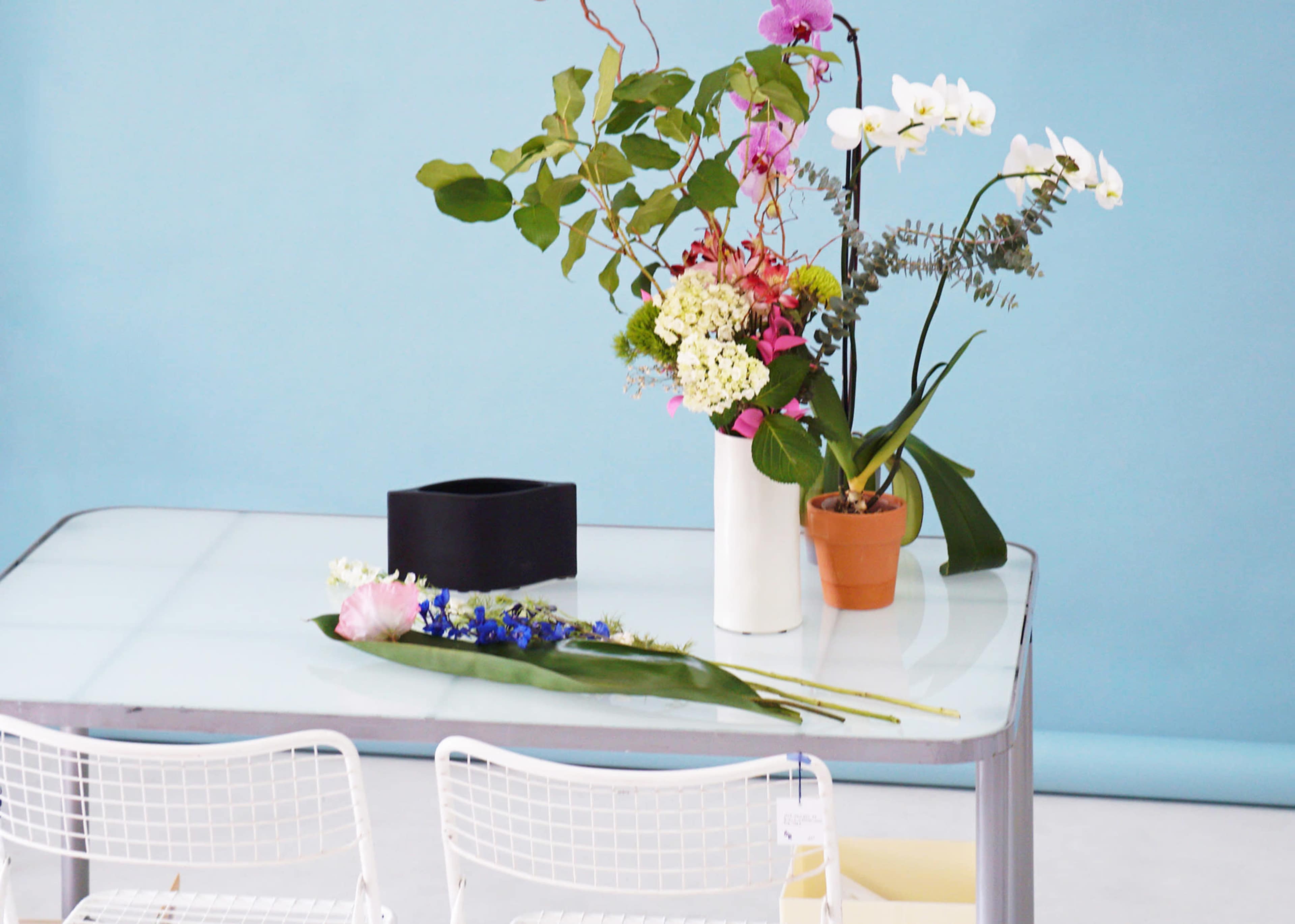 A glass table holds various flower arrangements and a potted plant against a light blue background.