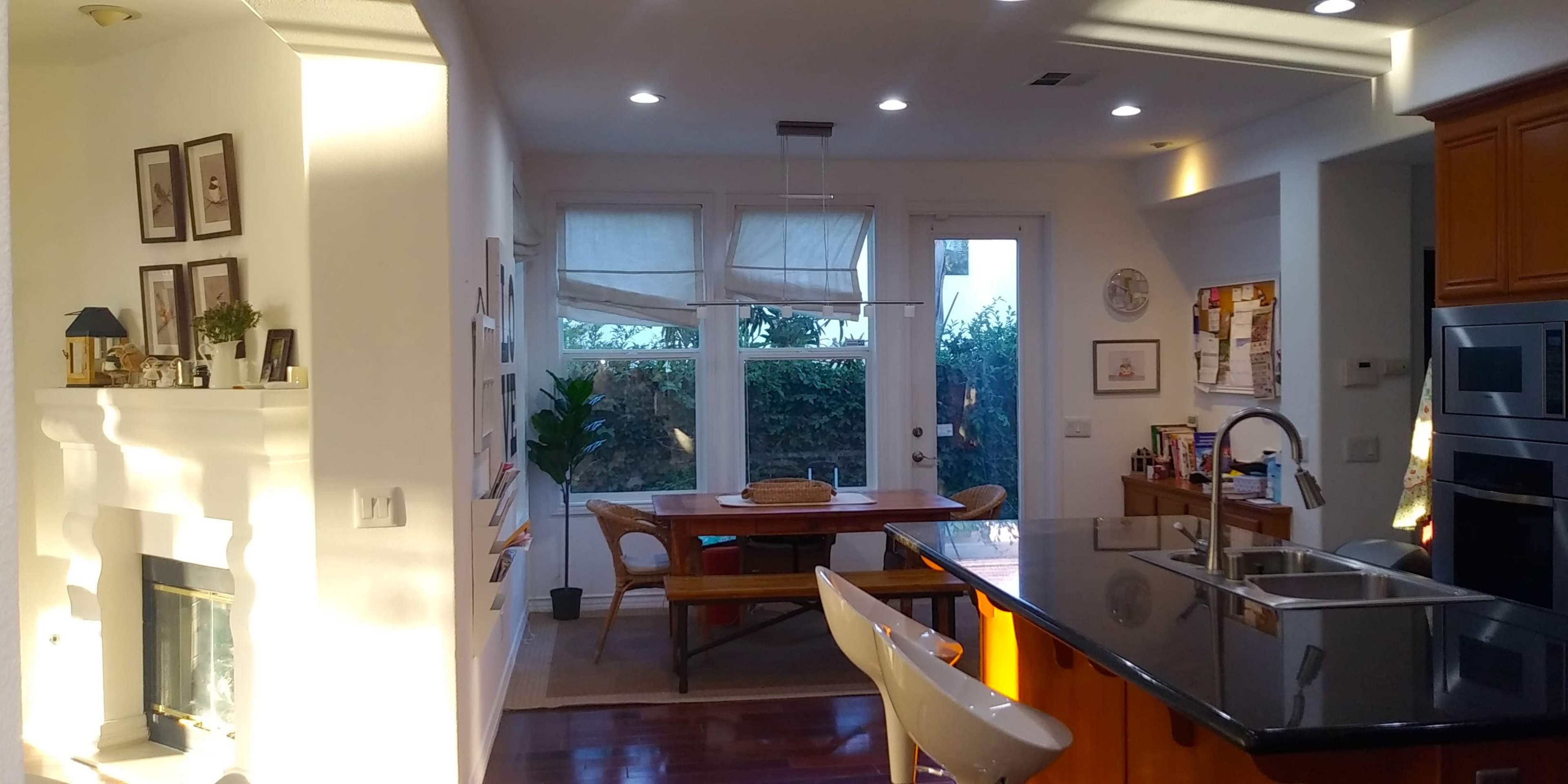 A modern kitchen with a black countertop, a dining area featuring a wooden table, and large windows allowing natural light to enter.