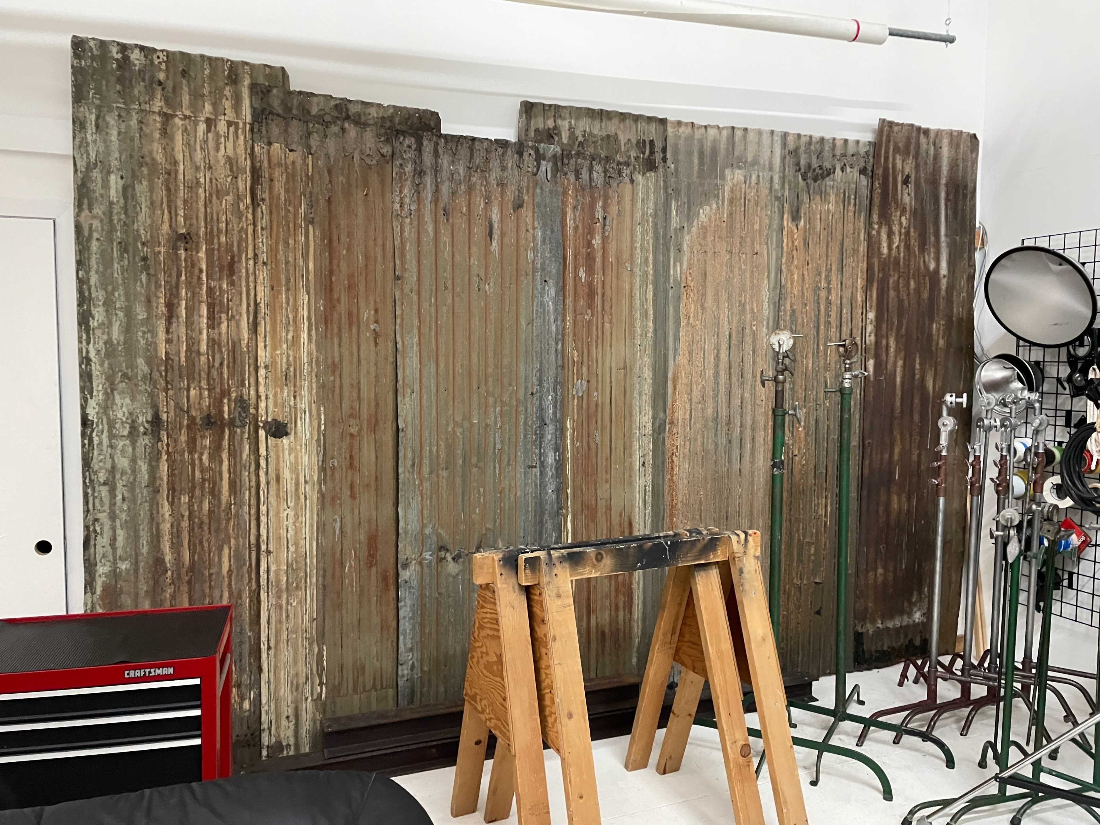 A weathered, corrugated metal backdrop leaning against a wall, accompanied by a wooden sawhorse and a red toolbox in a studio setting.