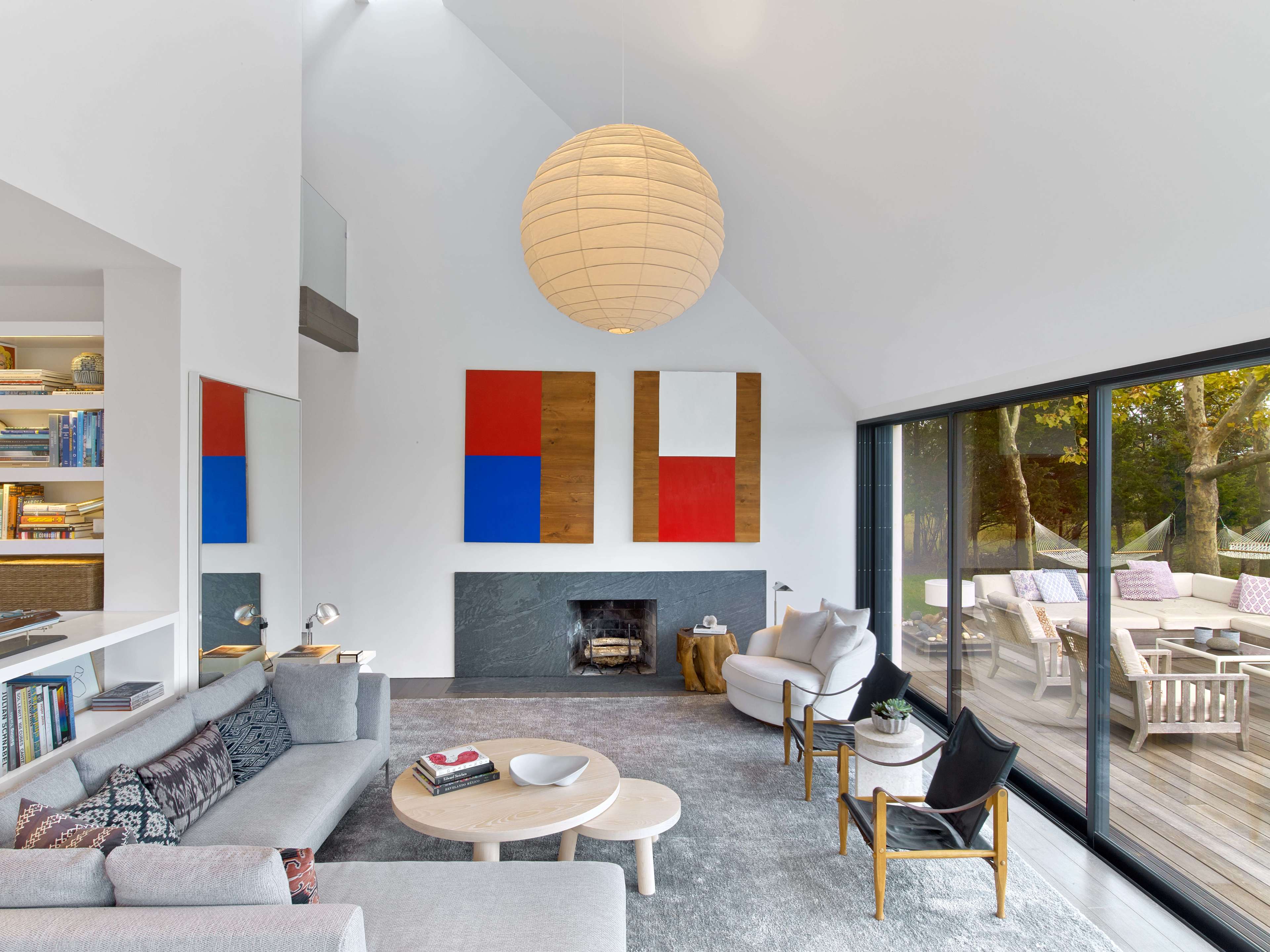 A modern living room features a prominent fireplace, two abstract paintings on the wall, and large glass doors opening to a deck surrounded by trees.