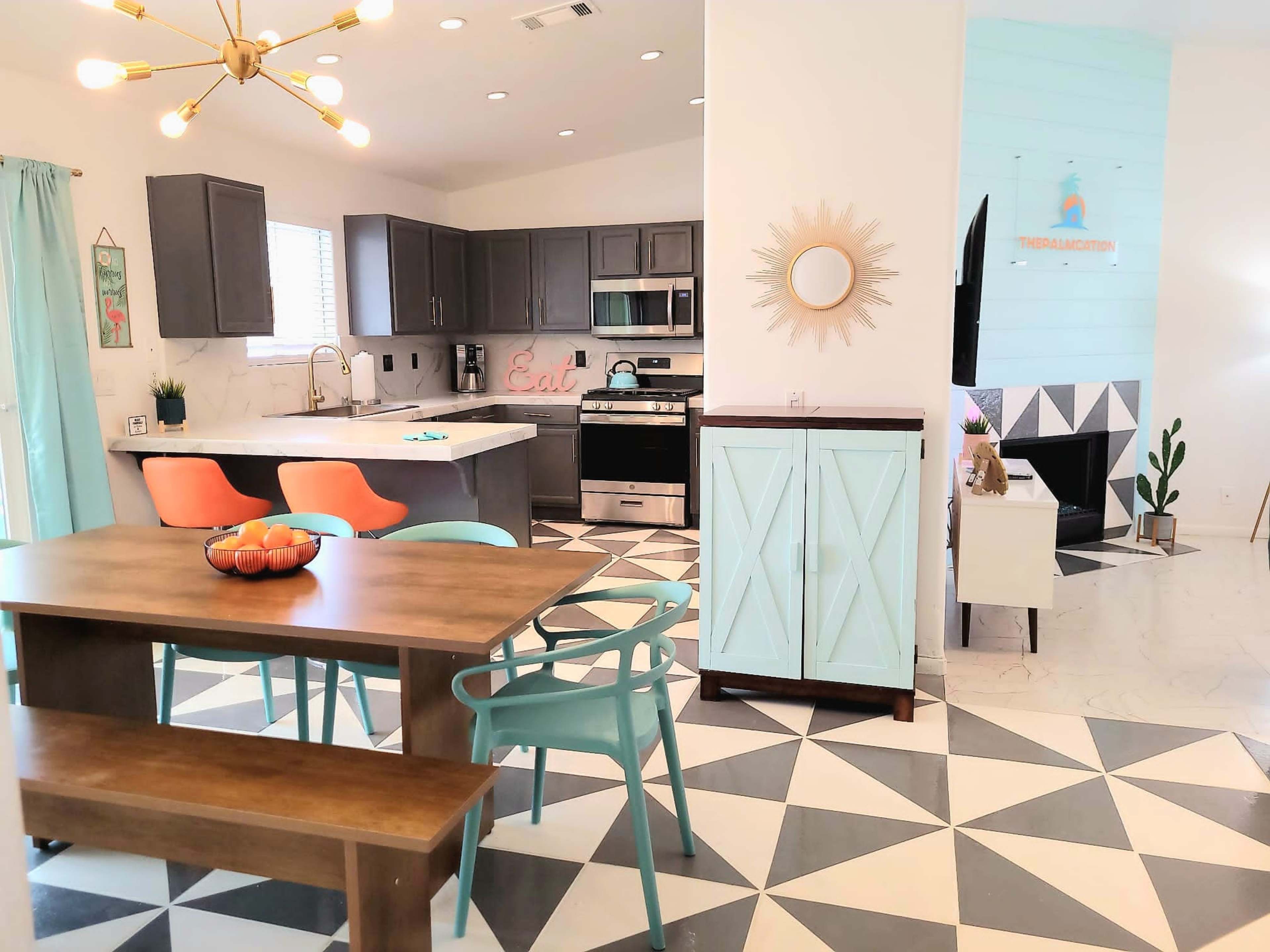 The image shows a modern kitchen and dining area featuring gray cabinetry, a wooden dining table, colorful chairs, and geometric patterned flooring.