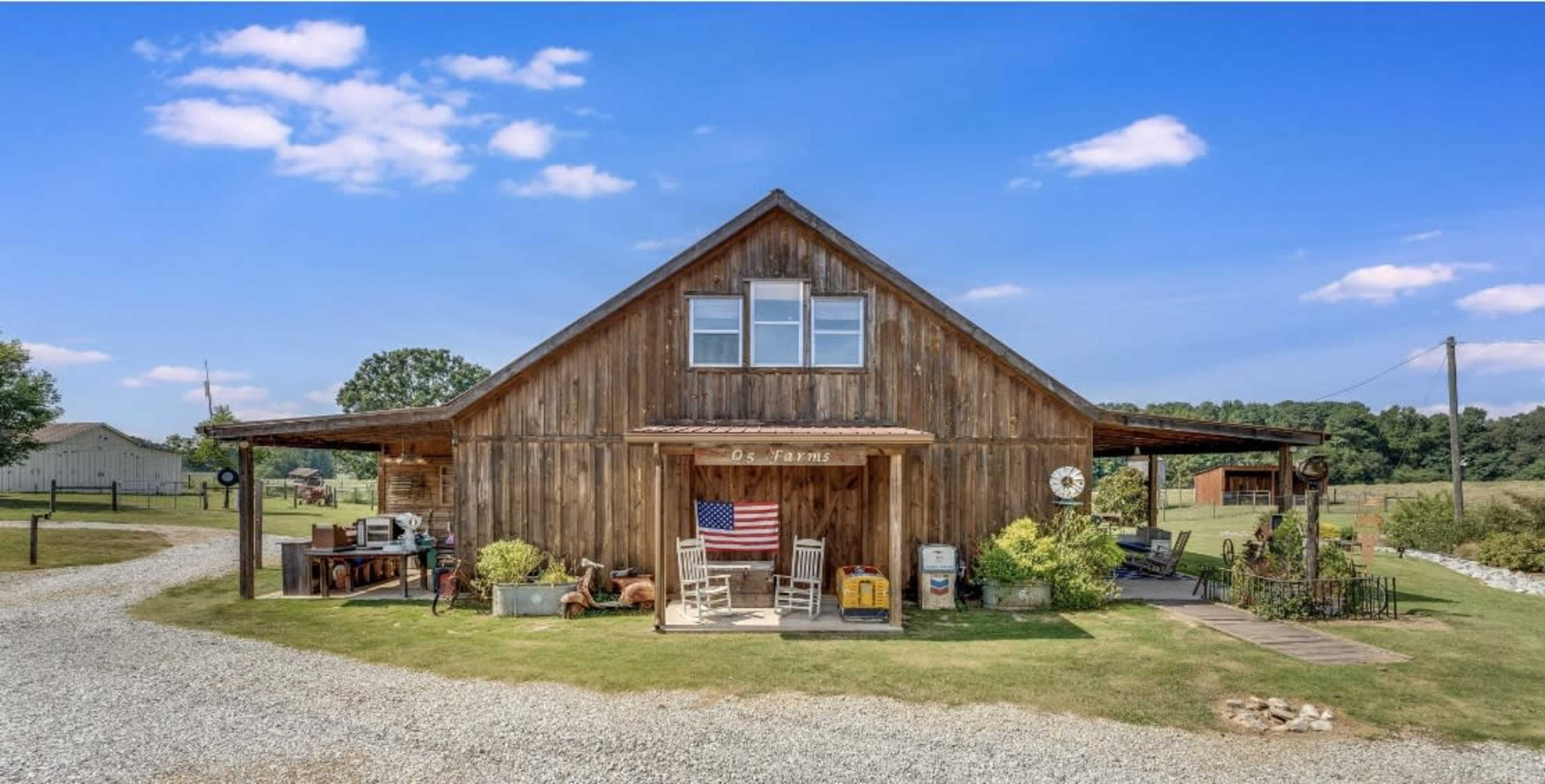A rustic wooden barn with an American flag displayed, surrounded by a gravel driveway and greenery under a clear blue sky.