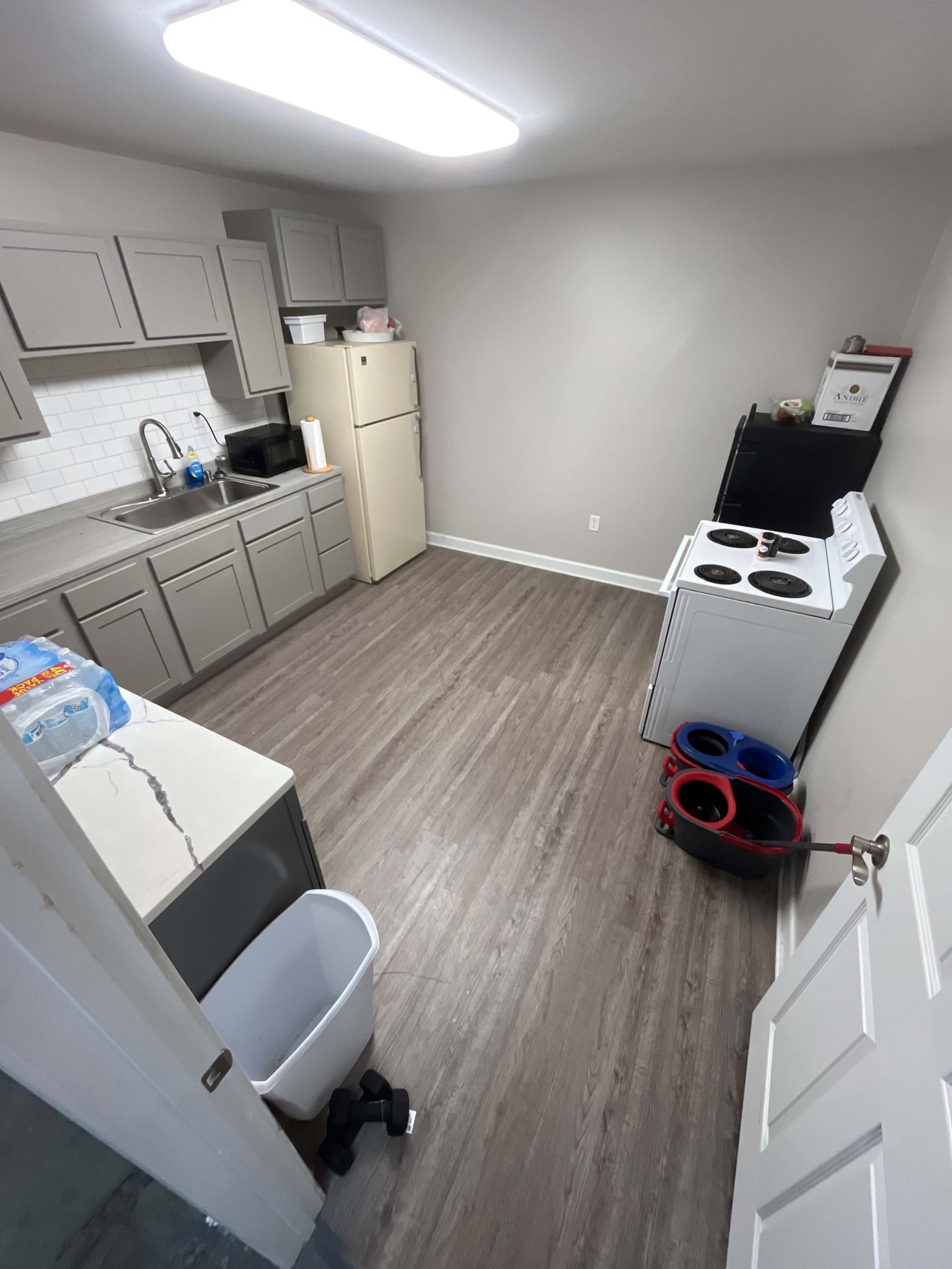 The image shows a compact kitchen with gray cabinets, a white refrigerator, a stove, and a sink, featuring a mix of appliances and a plastic trash can.