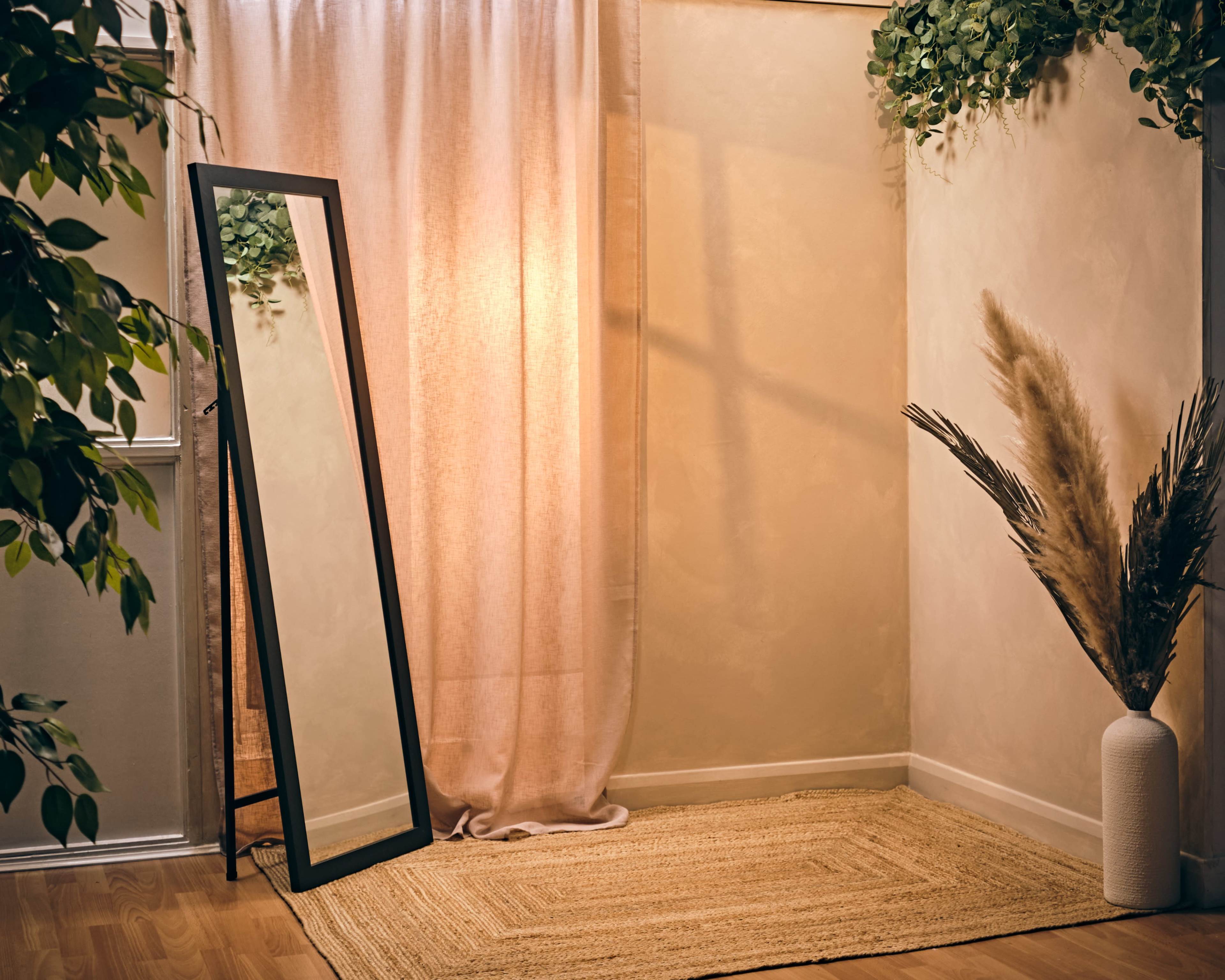 A full-length mirror stands beside a curtain in a softly lit corner, accented by a textured rug and a decorative plant arrangement.