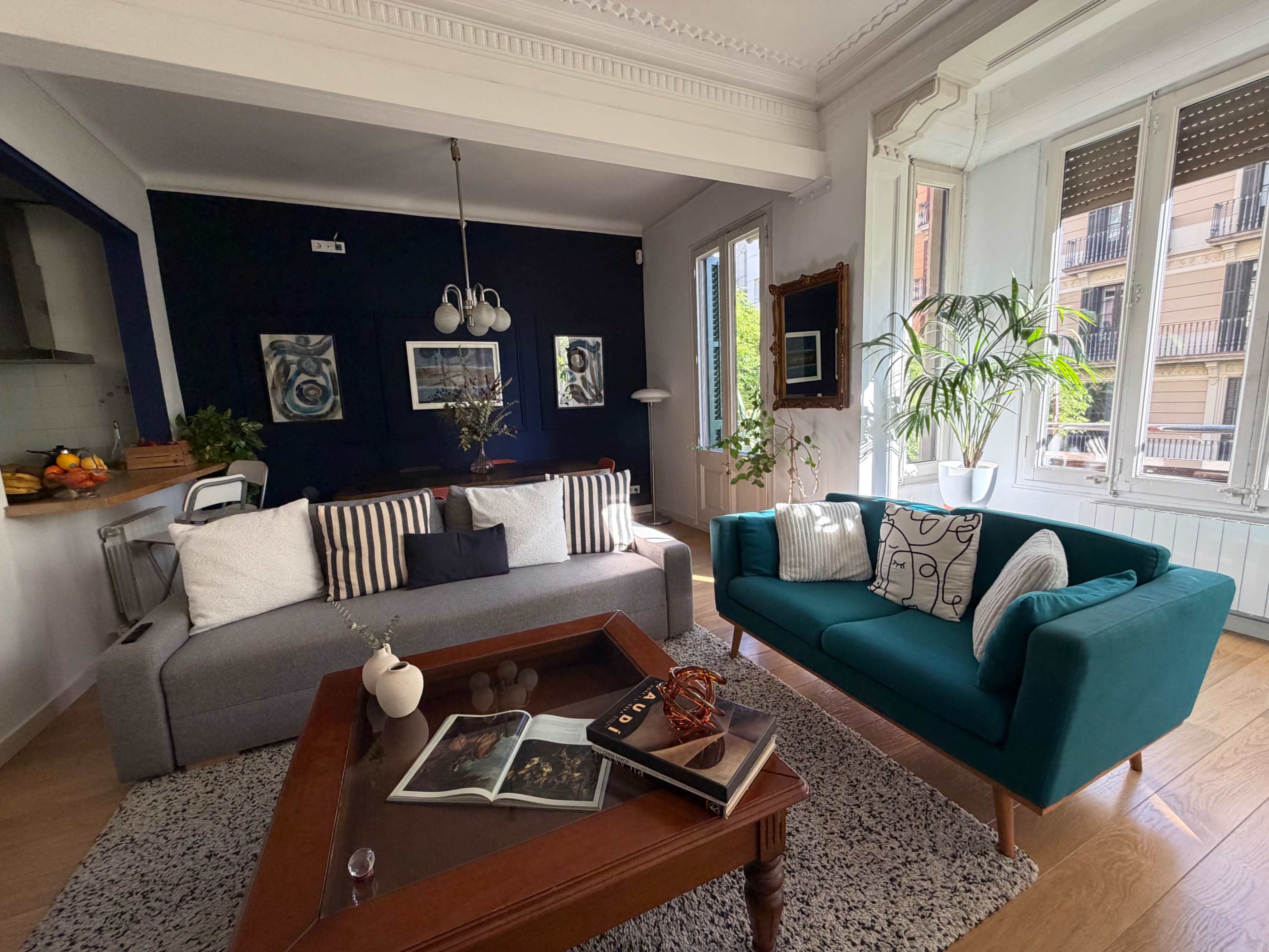 The image shows a modern living room featuring a gray sofa, a teal armchair, a wooden coffee table with books, and large windows allowing natural light to fill the space.