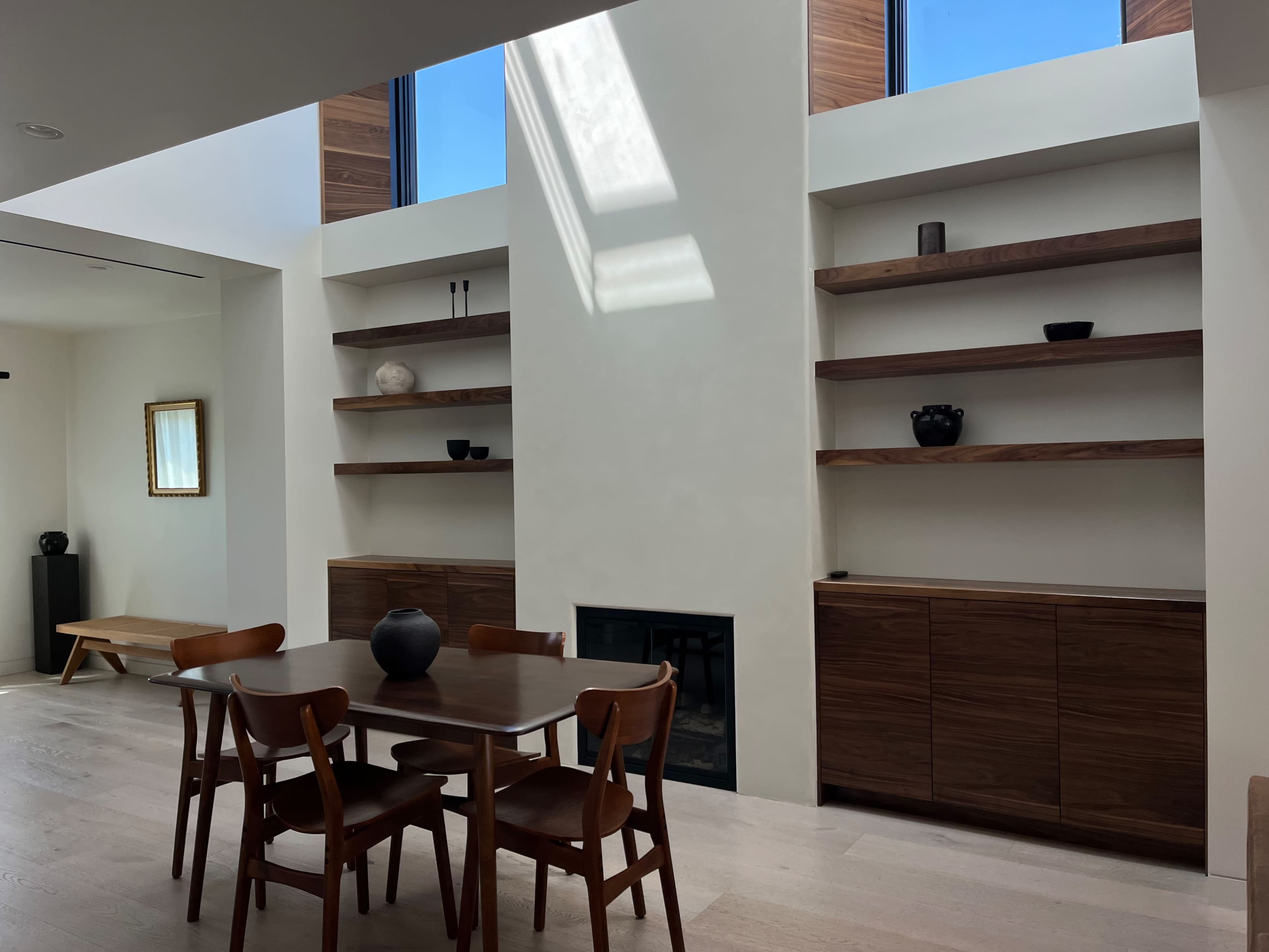 The image shows a modern interior featuring a dining table with chairs, built-in shelves, and large windows above.