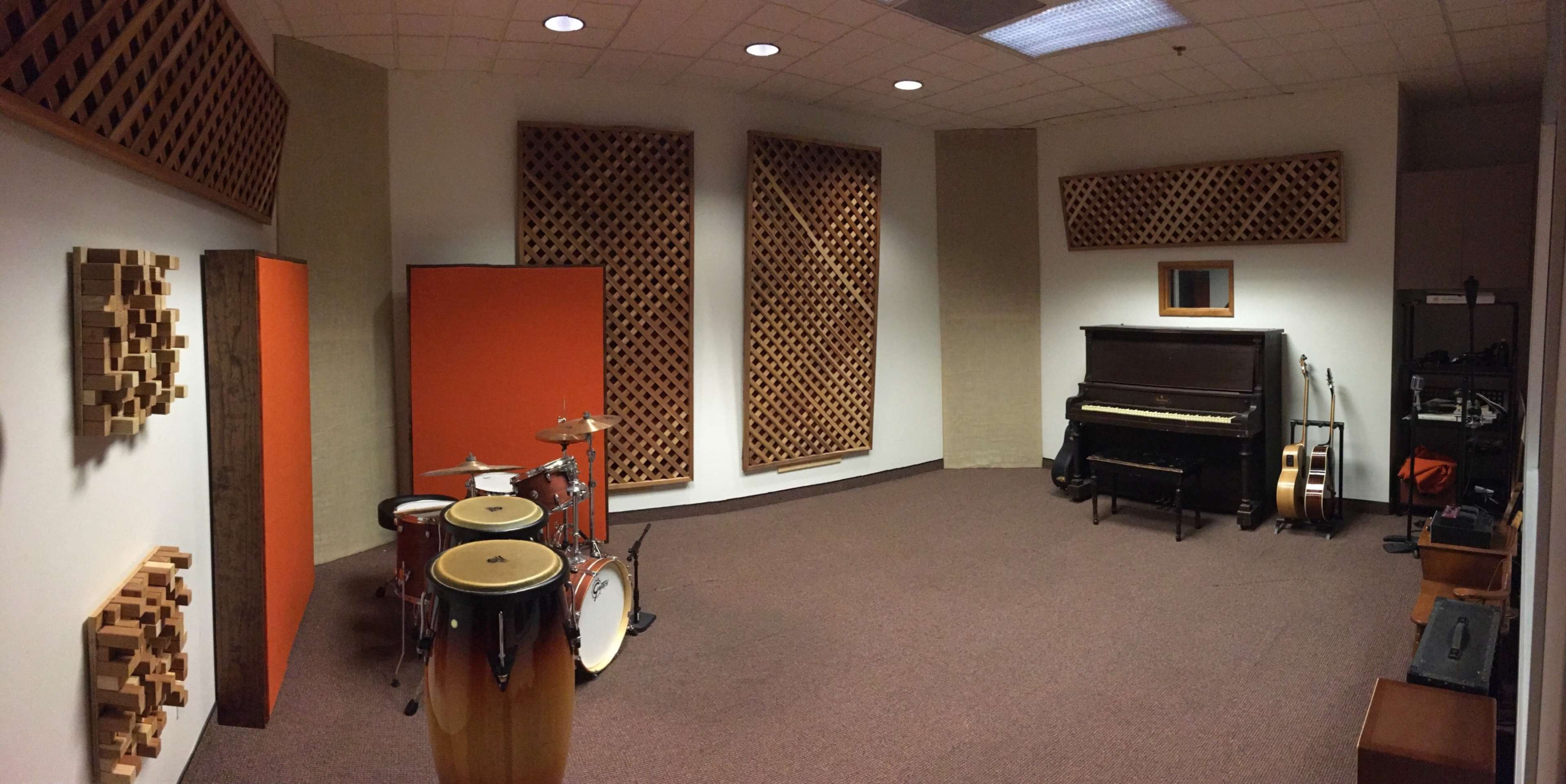 A music rehearsal room with various instruments, including a drum set, congas, a piano, and guitars, surrounded by acoustic panels on the walls.