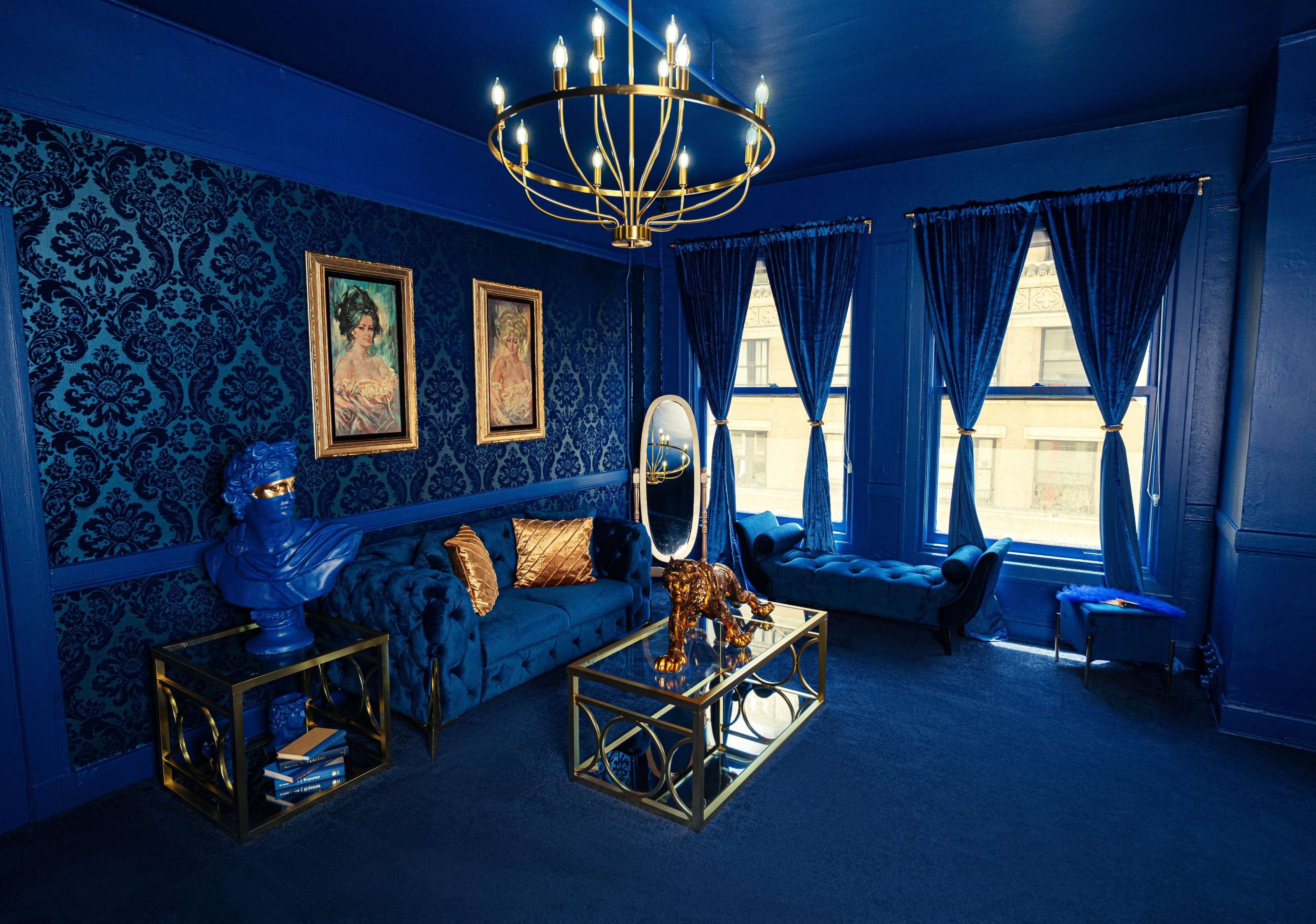 The room features blue walls with patterned wallpaper, a blue velvet sofa, and a large chandelier hanging from the ceiling.