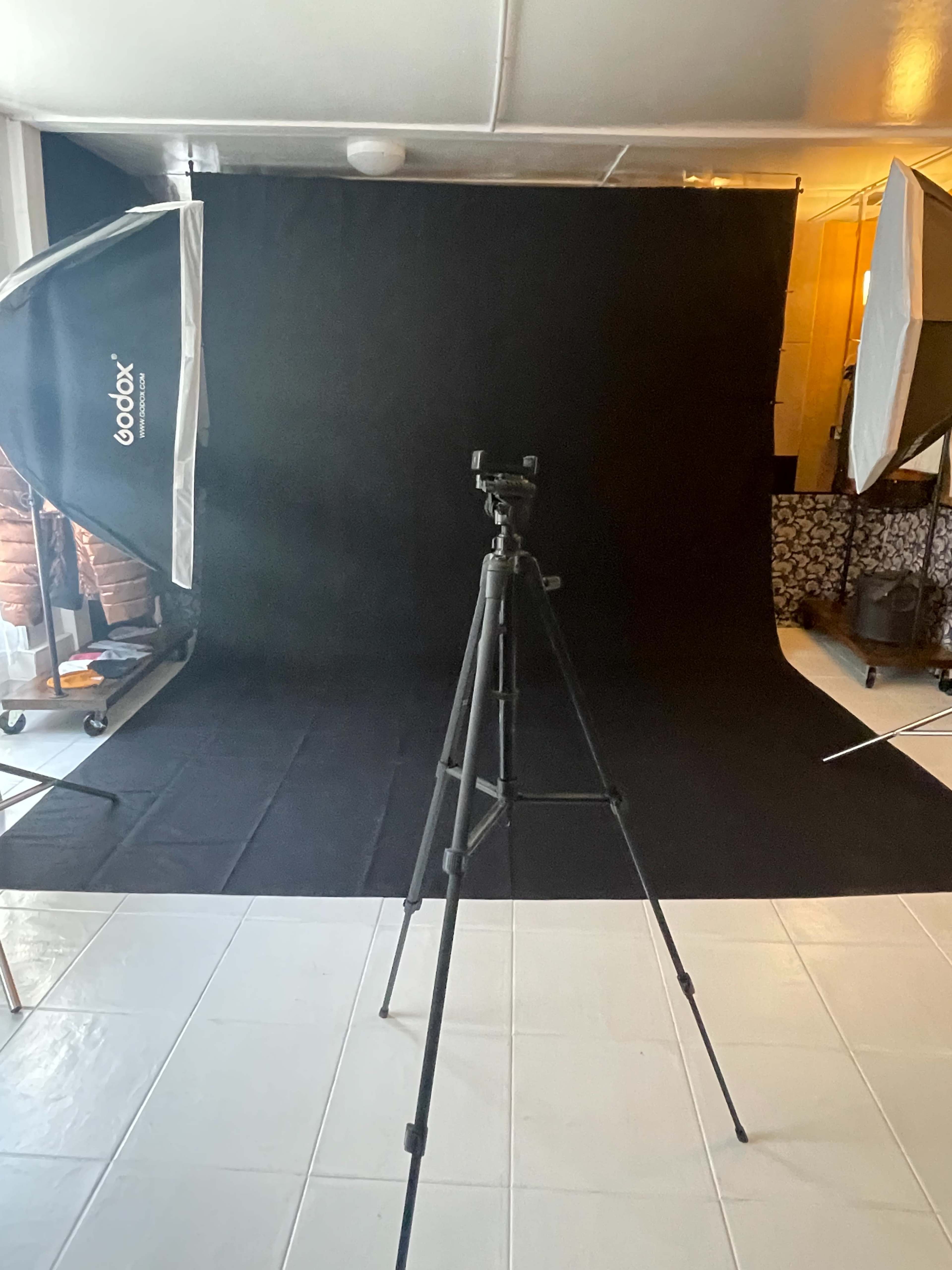 The image shows a photography studio setup with a black backdrop, a camera tripod, and two softbox light stands on either side.