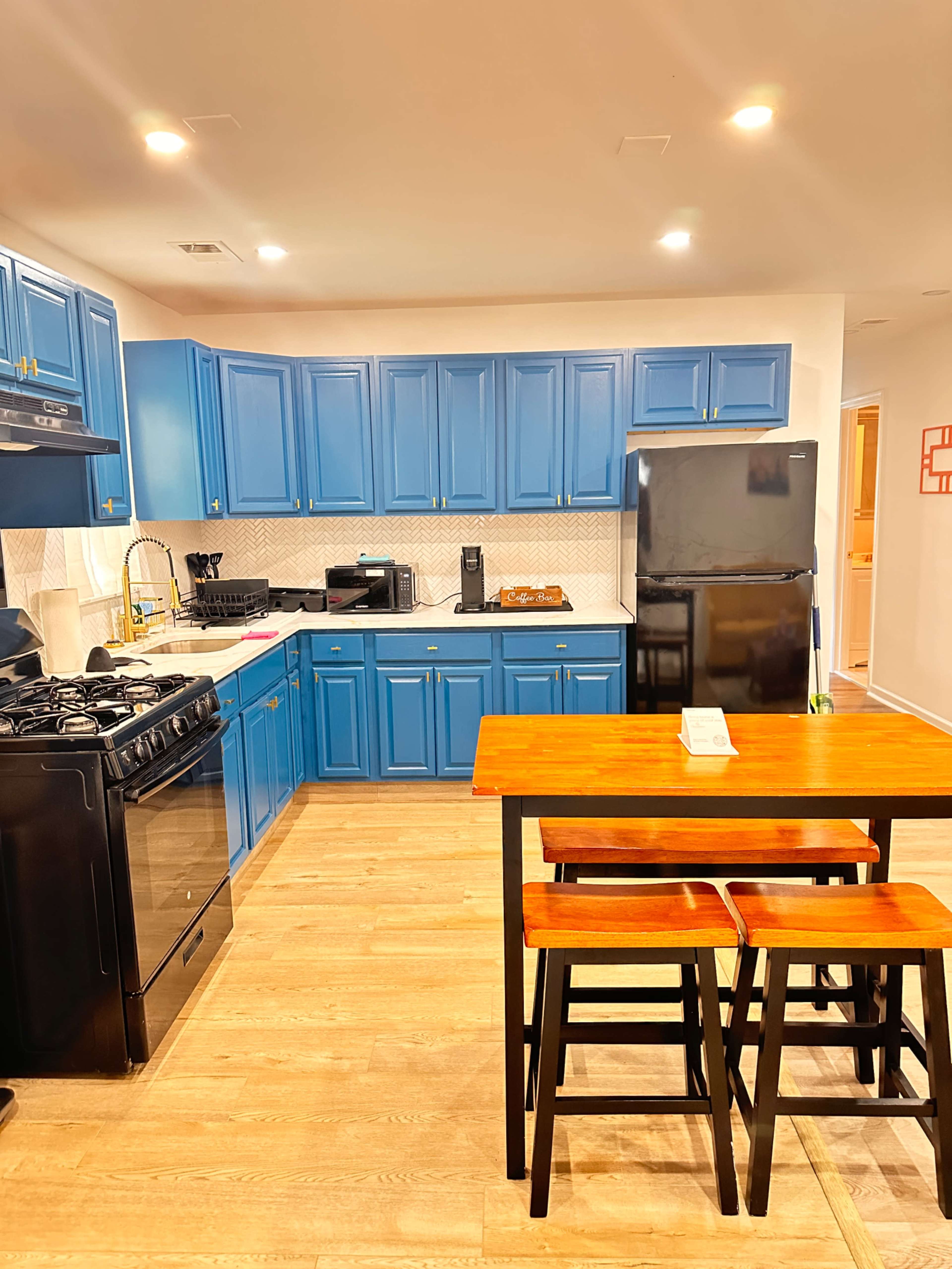 The image shows a modern kitchen featuring bright blue cabinetry, a black refrigerator, a stove, and a small wooden dining table with three stools.