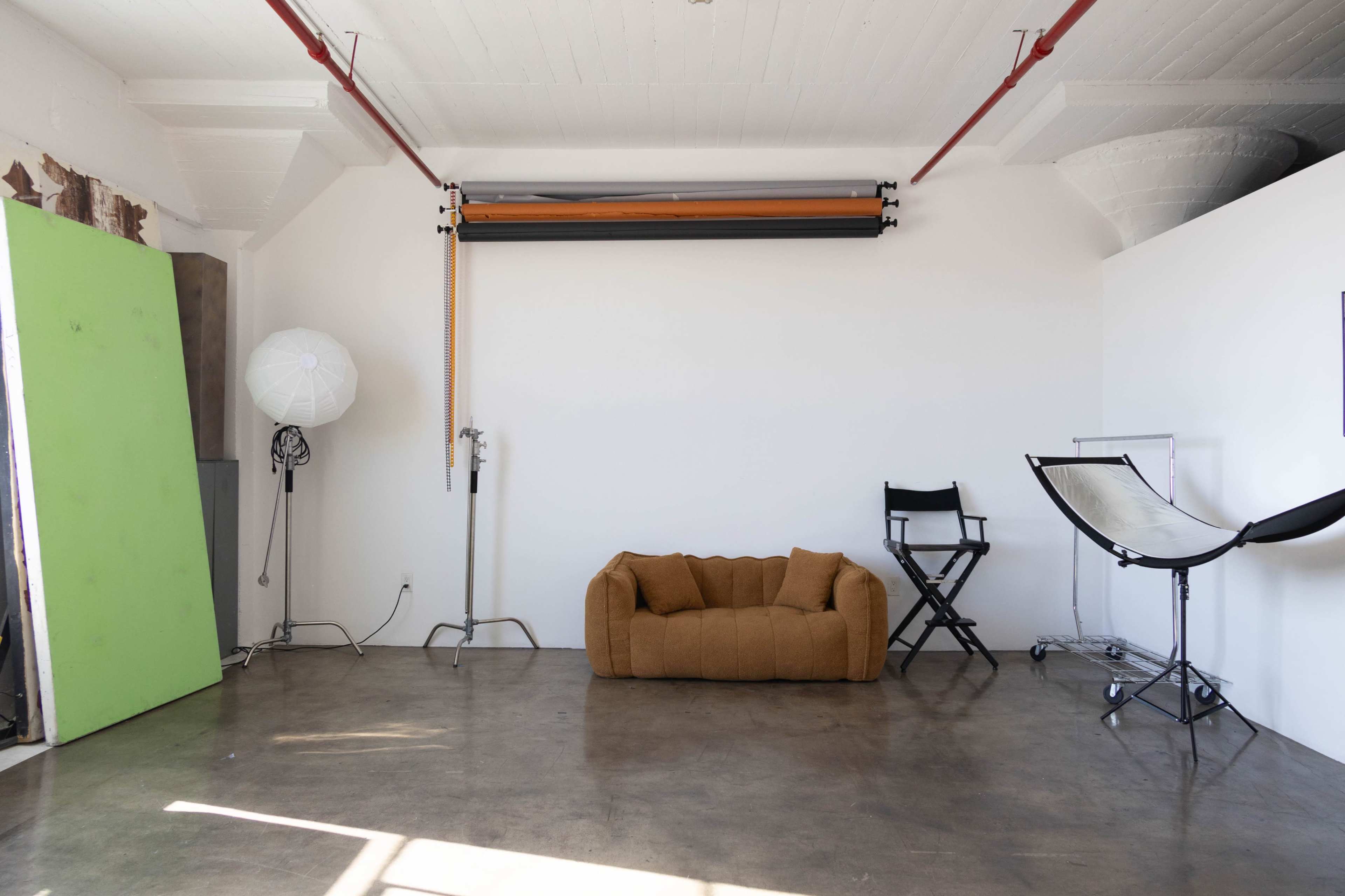 The image shows a spacious, minimalist studio with a brown couch, a director's chair, and lighting equipment against a white wall.