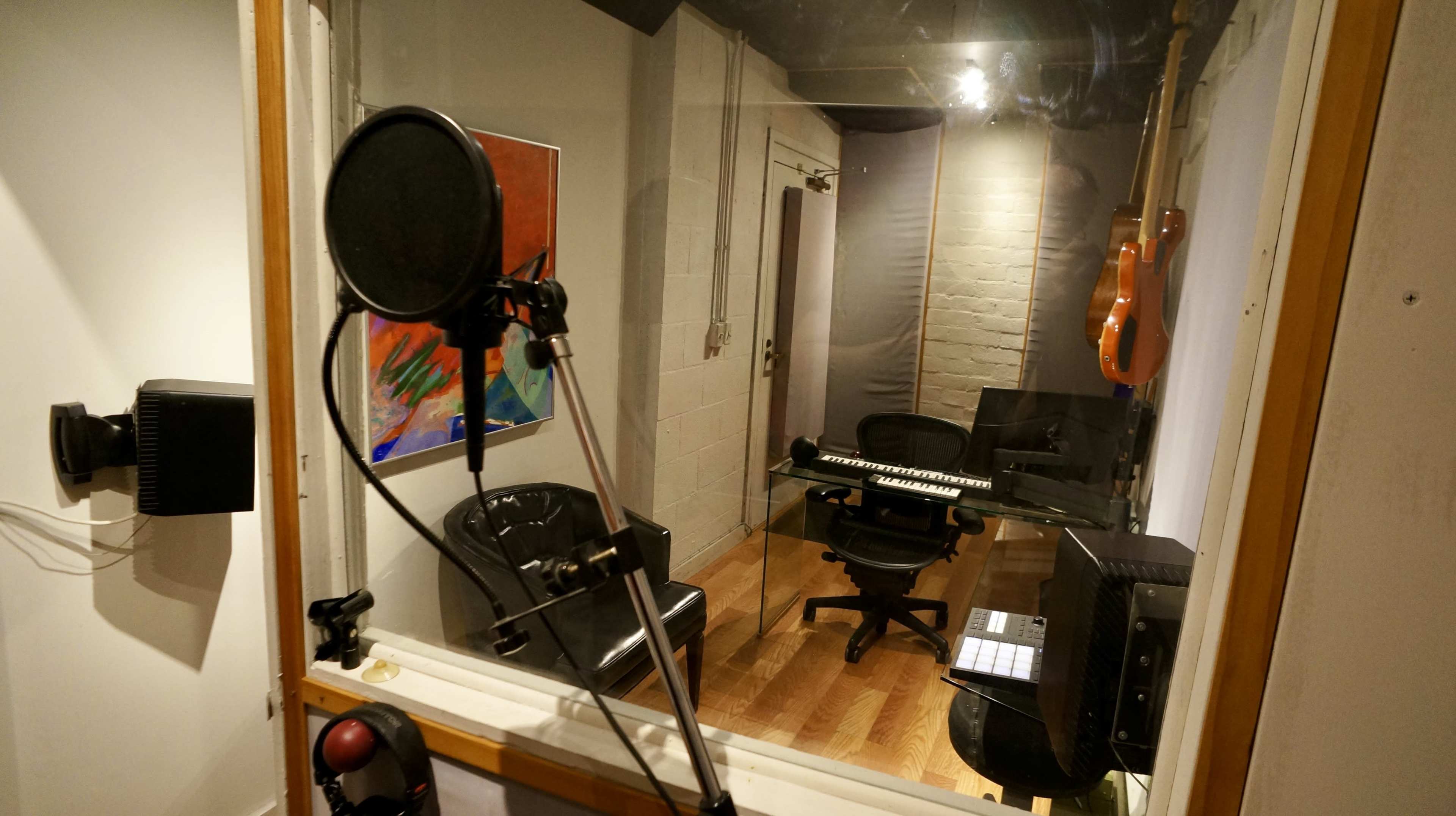 The image shows a small music recording studio with a microphone stand, a glass wall, an electric guitar hanging, and various instruments and chairs inside.