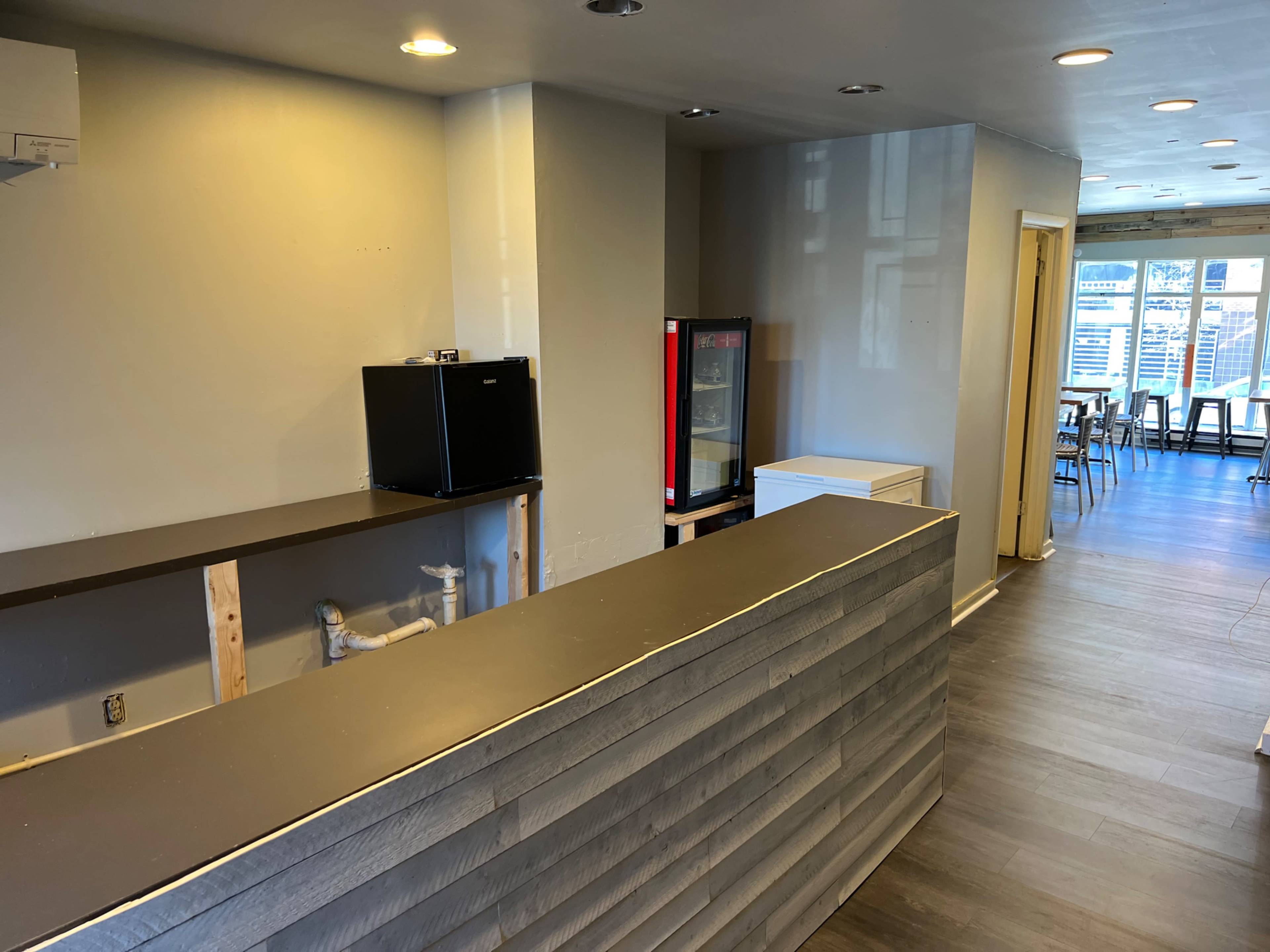A partially completed interior of a cafe with a counter, sink, and cooling units, set against a backdrop of modern flooring and visible seating.