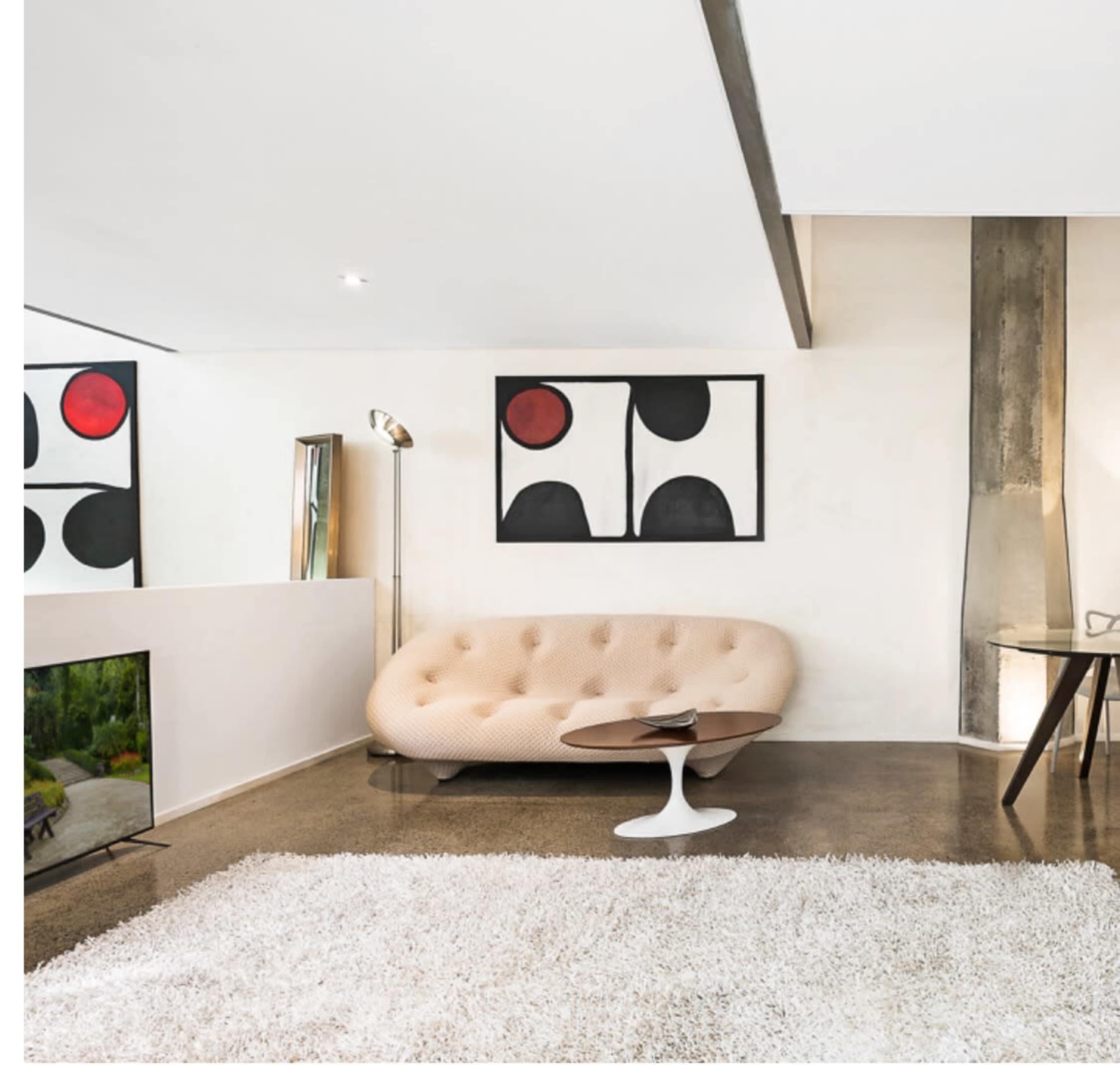 The image shows a modern living room with a rounded beige sofa, abstract wall art, a small coffee table, and a sleek television on a low cabinet, featuring polished concrete flooring and a white rug.