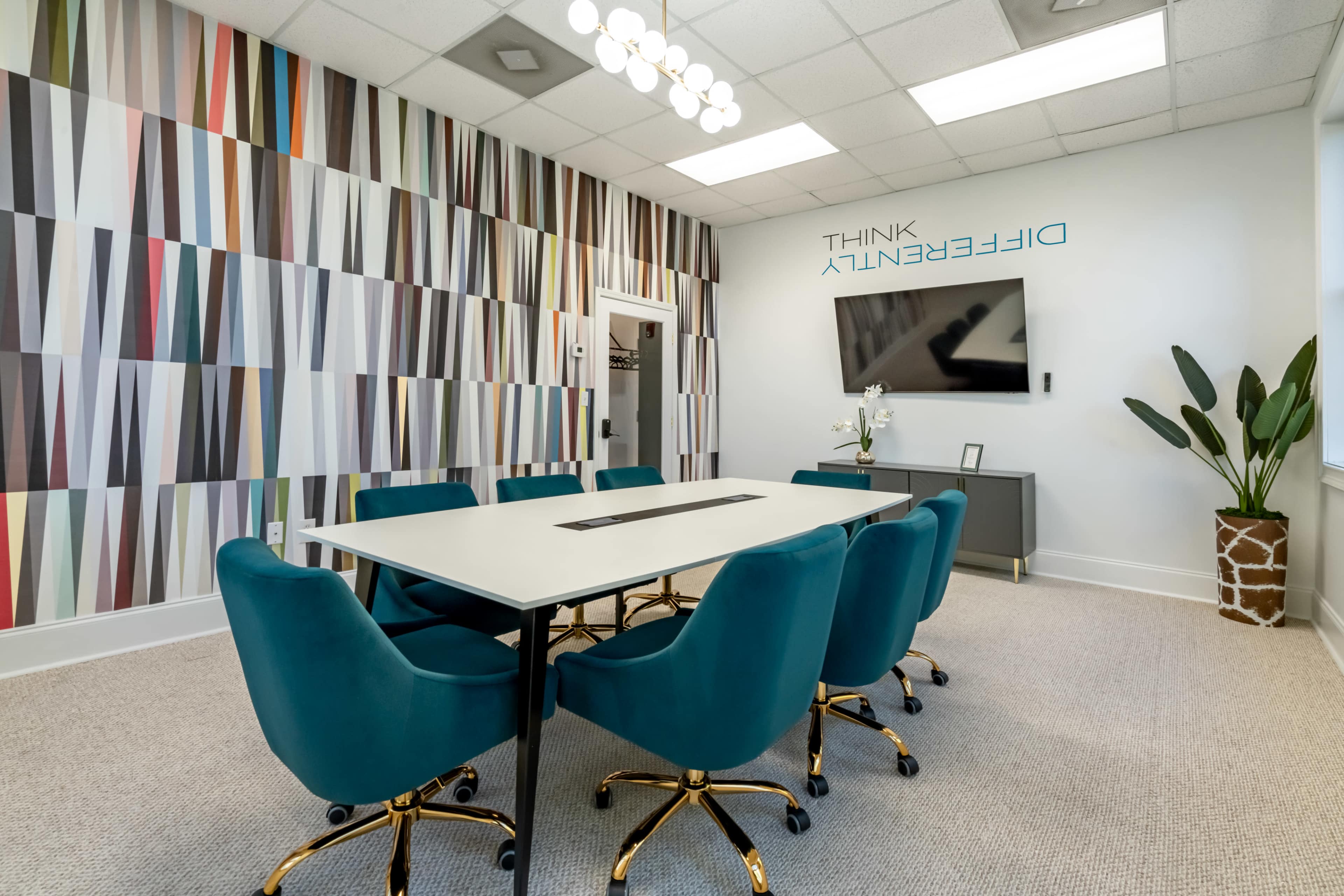 A modern conference room features a large rectangular table surrounded by teal chairs and a wall with colorful geometric artwork.