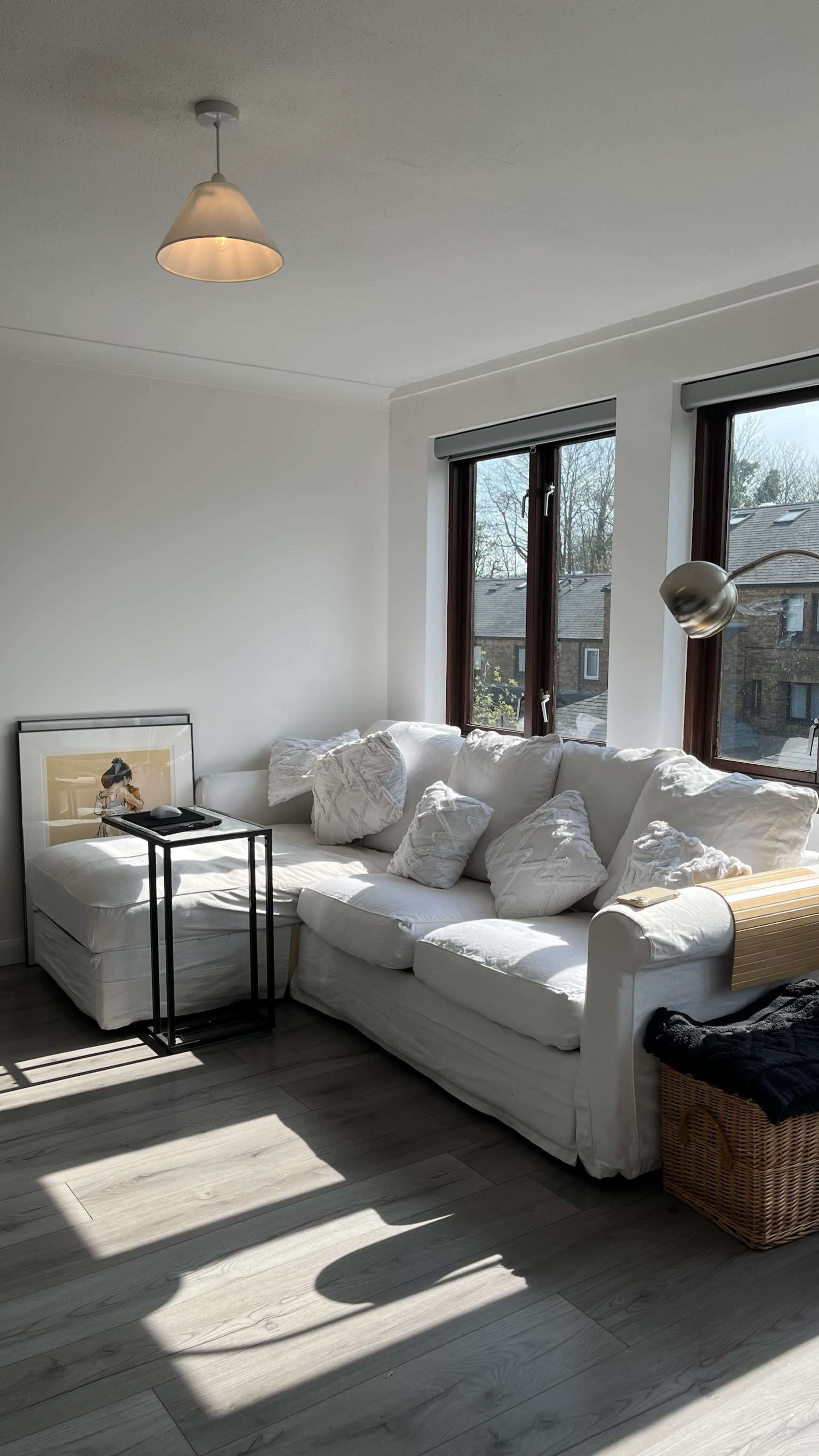 A bright living room features a white sofa adorned with plush cushions, a small black side table, and large windows allowing ample natural light.