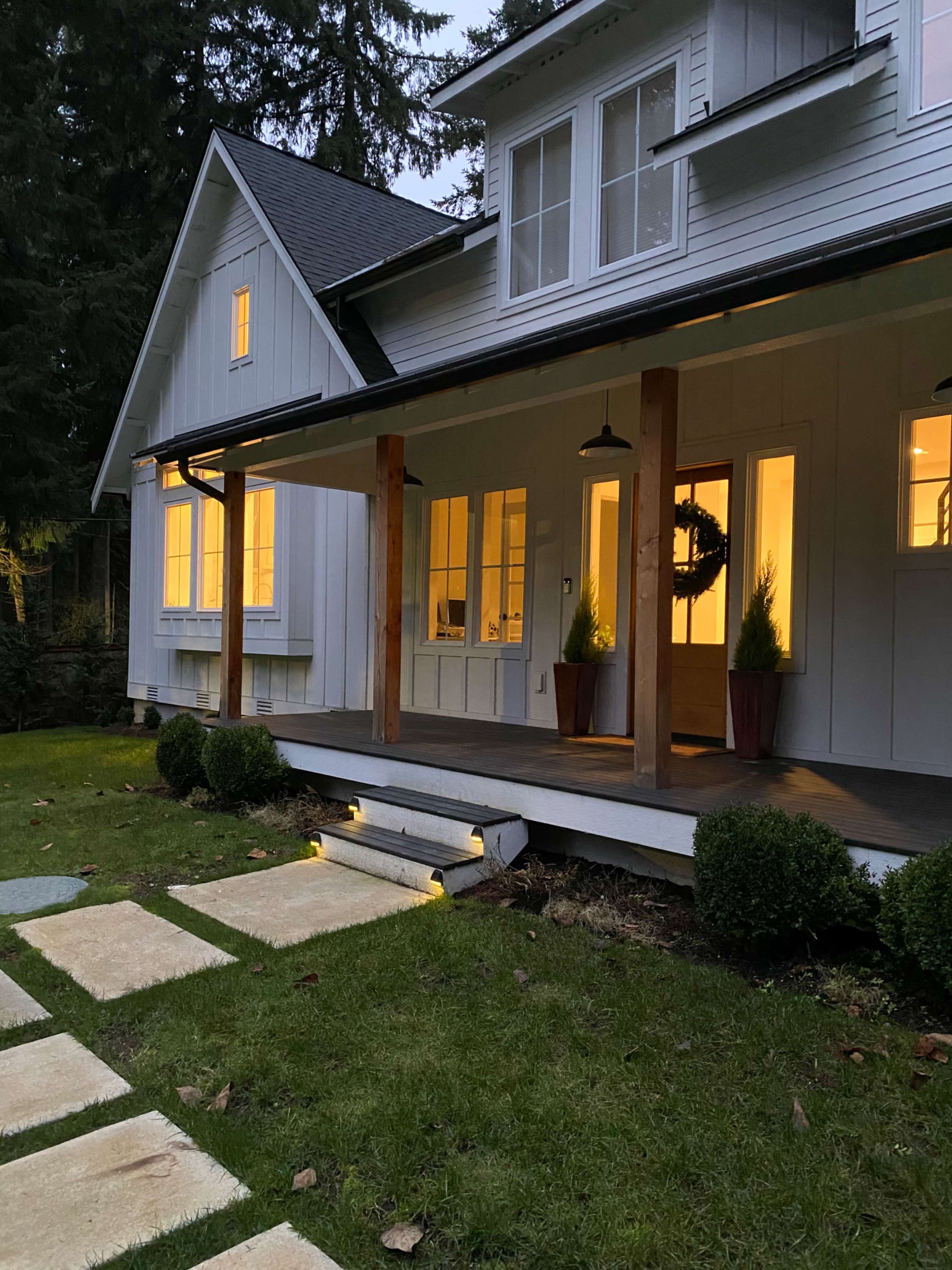 A modern house features a porch with multiple large windows, two potted plants, and a stone pathway leading to the entrance, surrounded by grass and trees.