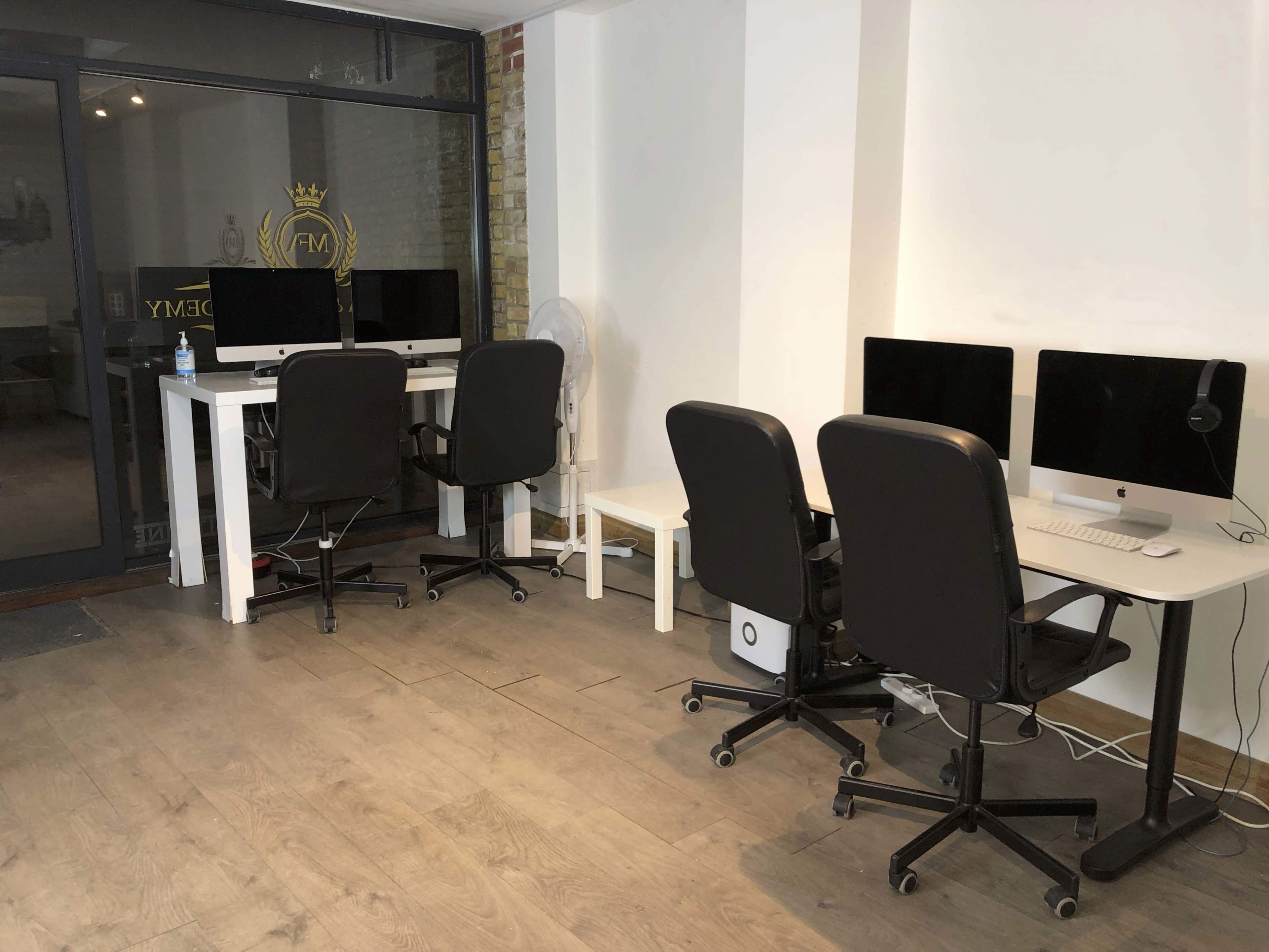 The image shows a workspace with two desks featuring desktop computers and three black rolling chairs arranged against a white wall.