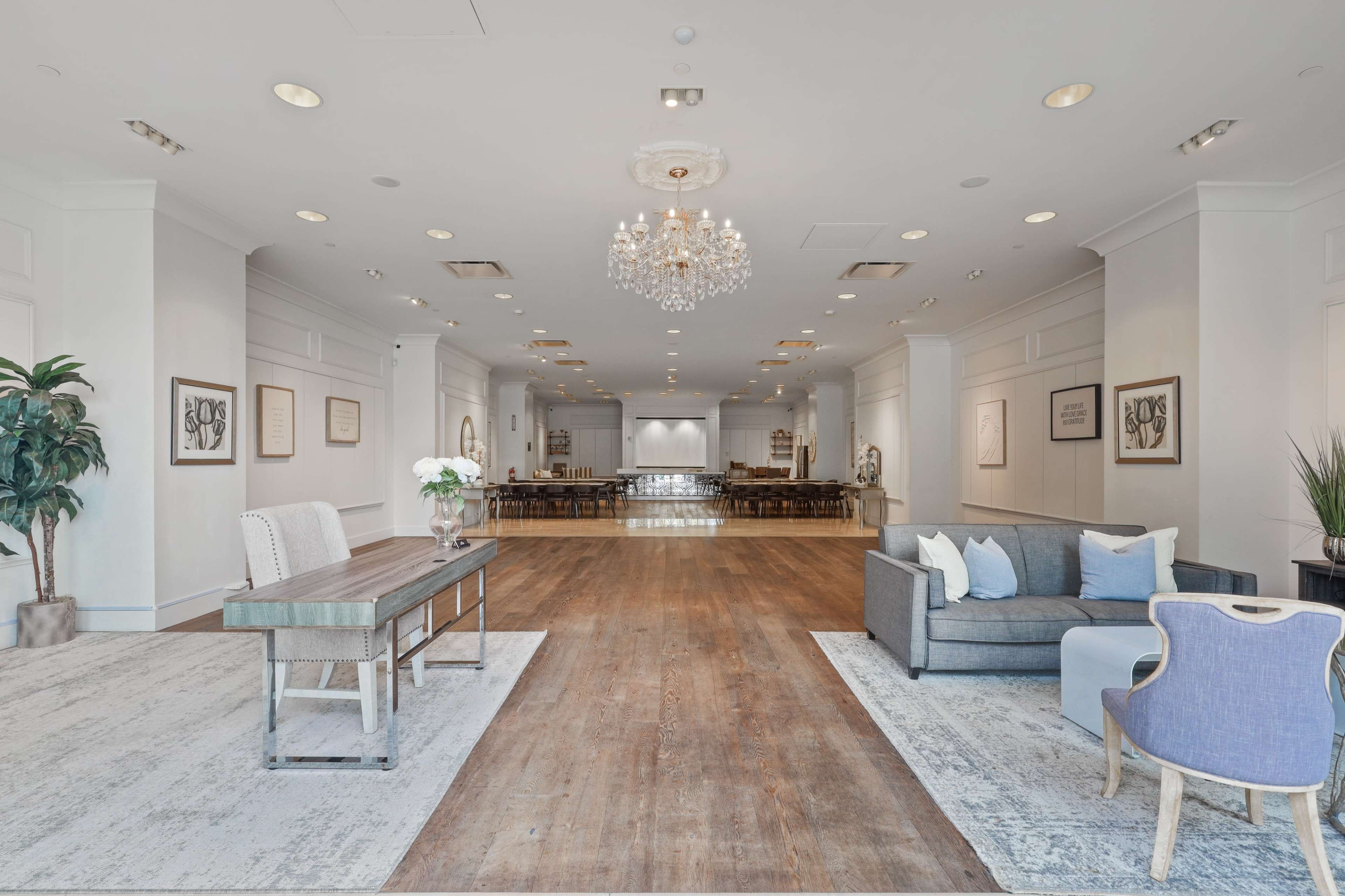 The image shows a spacious event room featuring a large chandelier, wooden flooring, and seating arrangements with tables and chairs in a neutral color palette.