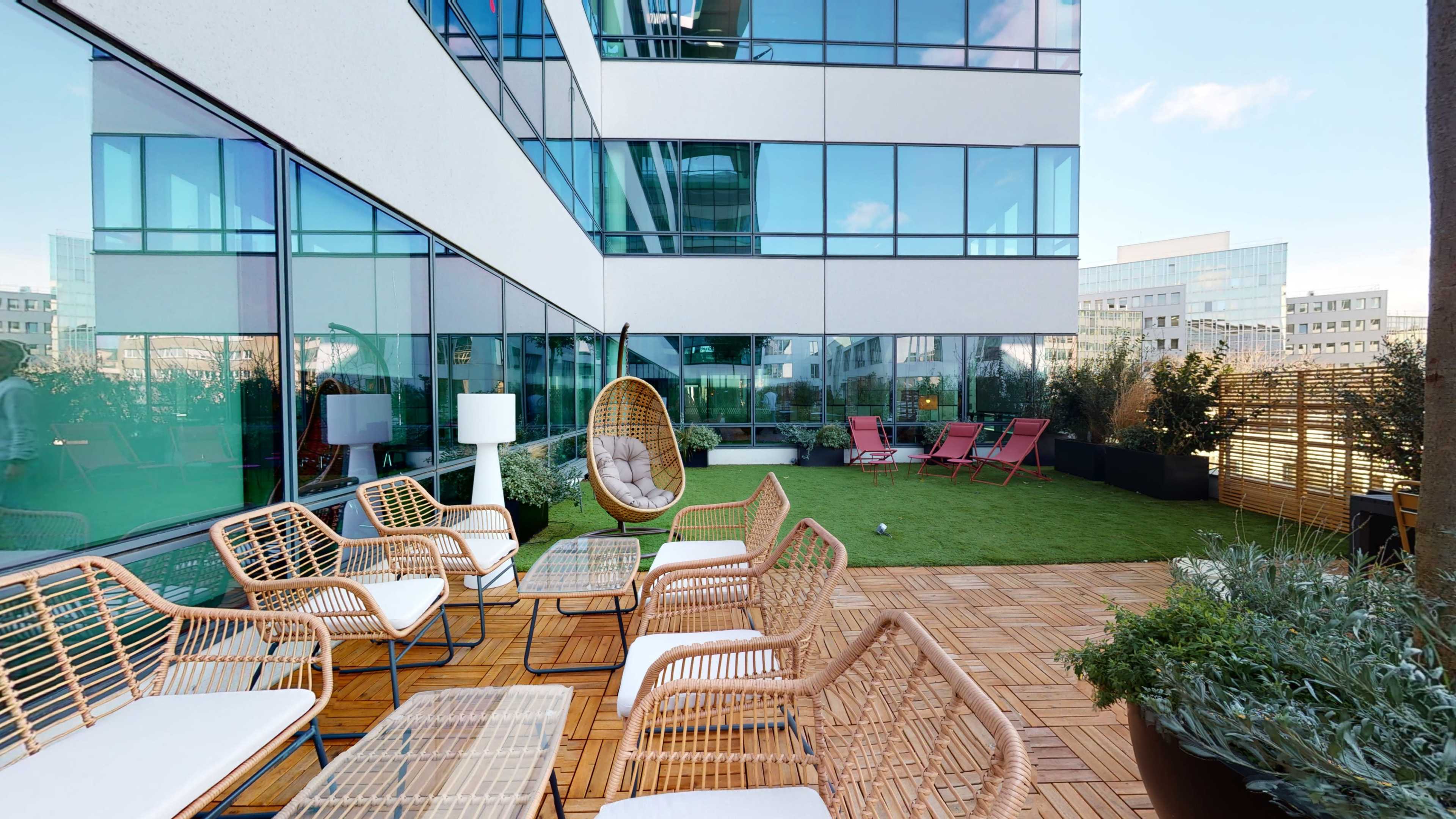 An outdoor lounge area with rattan furniture, including chairs and tables, on a wooden deck surrounded by greenery and modern glass buildings.