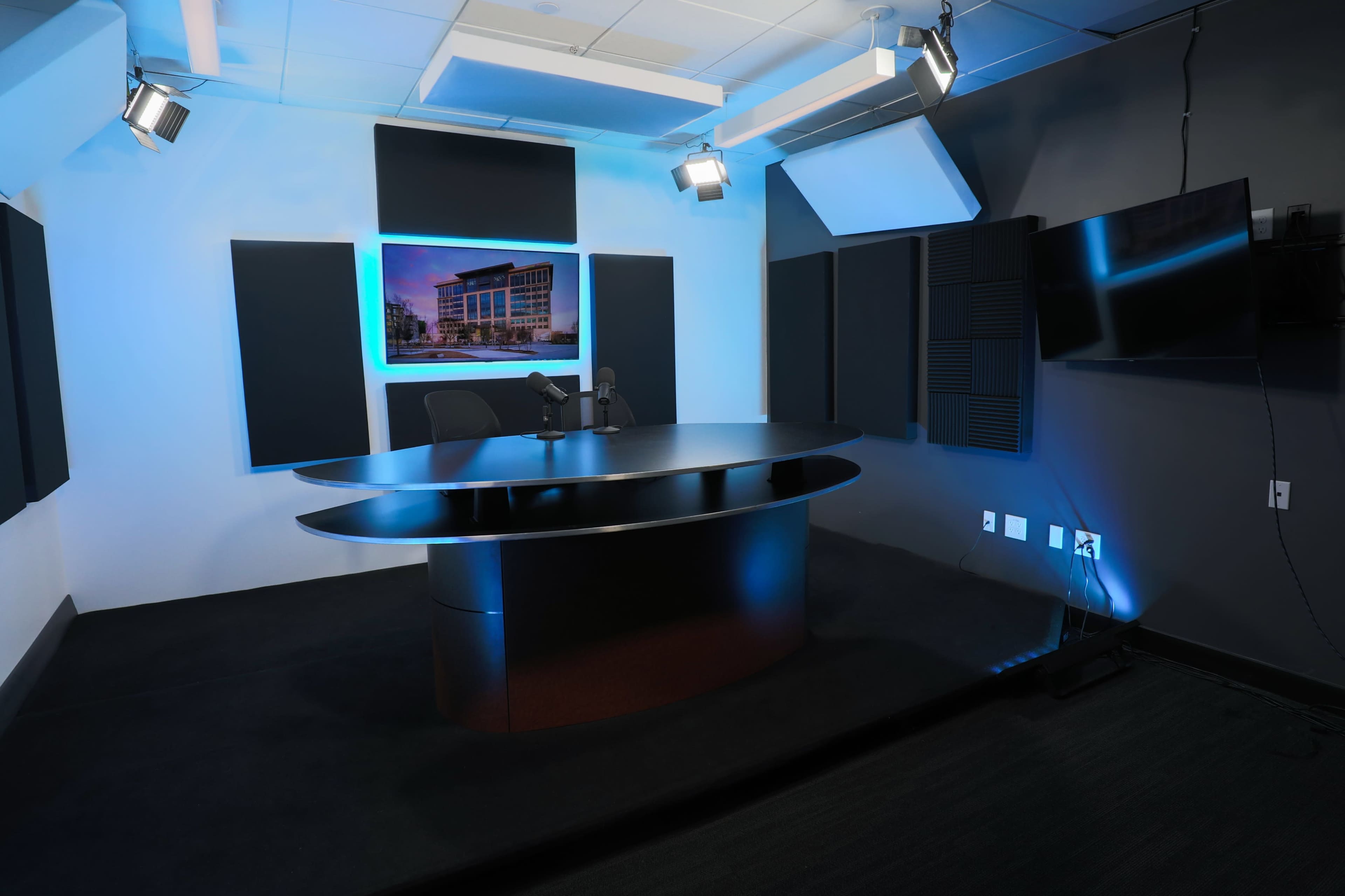 A modern broadcast studio featuring a sleek black desk, two chairs, and wall-mounted lighting, with a large screen displaying a building in the background.