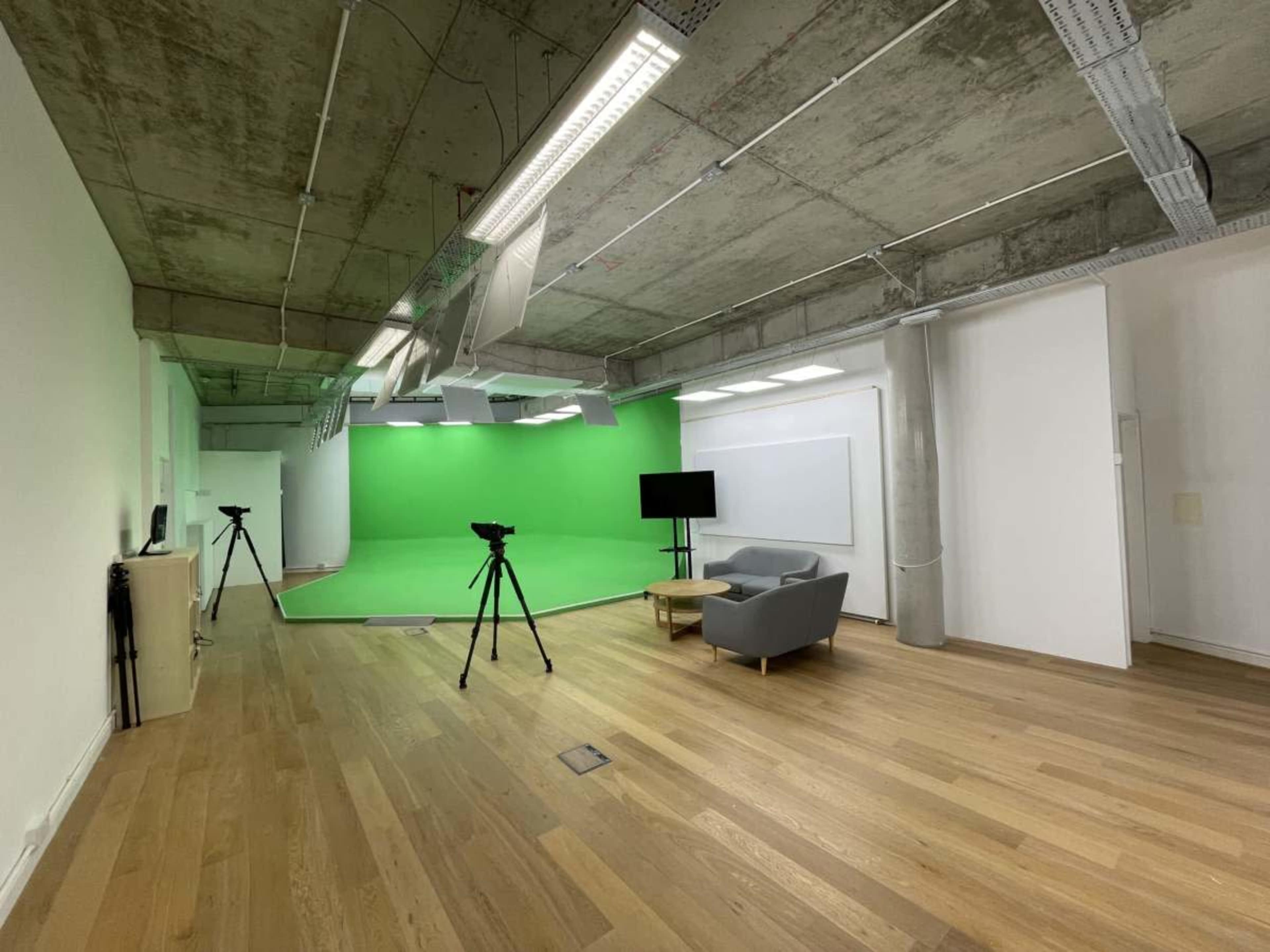 The image shows a minimalist studio space featuring a green screen backdrop, two cameras on tripods, a television, and a gray sofa on a wooden floor.