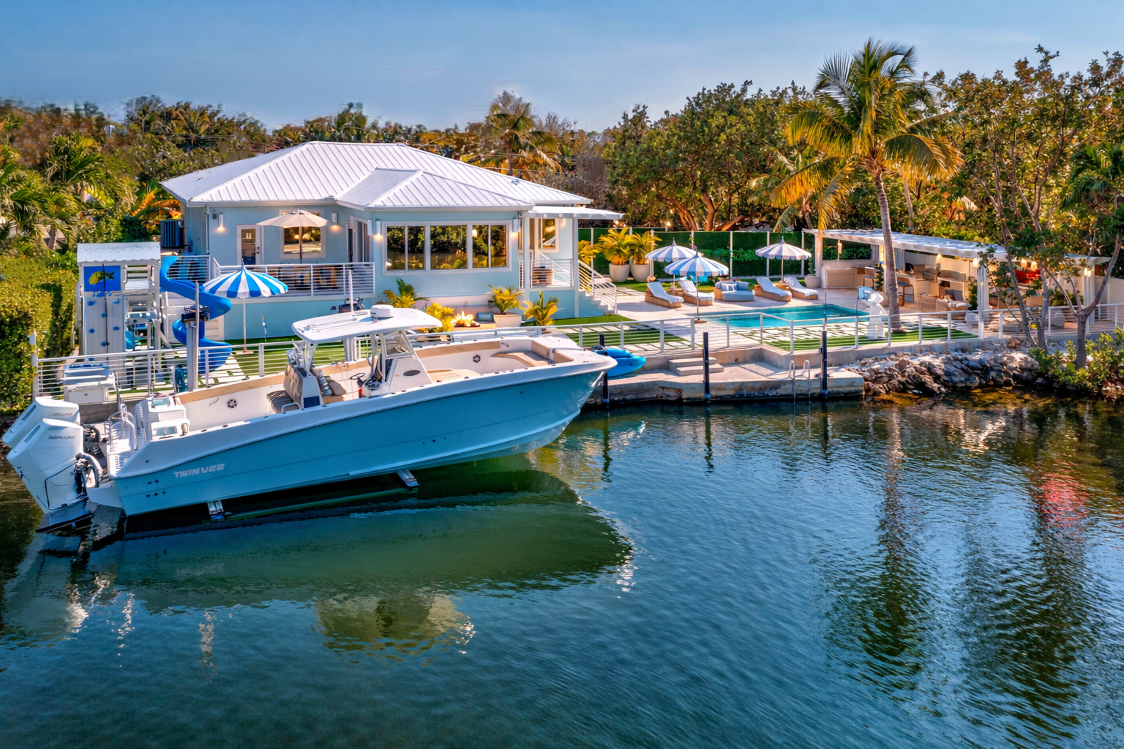 A modern waterfront house with a boat docked in front features a spacious deck with umbrellas and loungers overlooking a calm waterway.