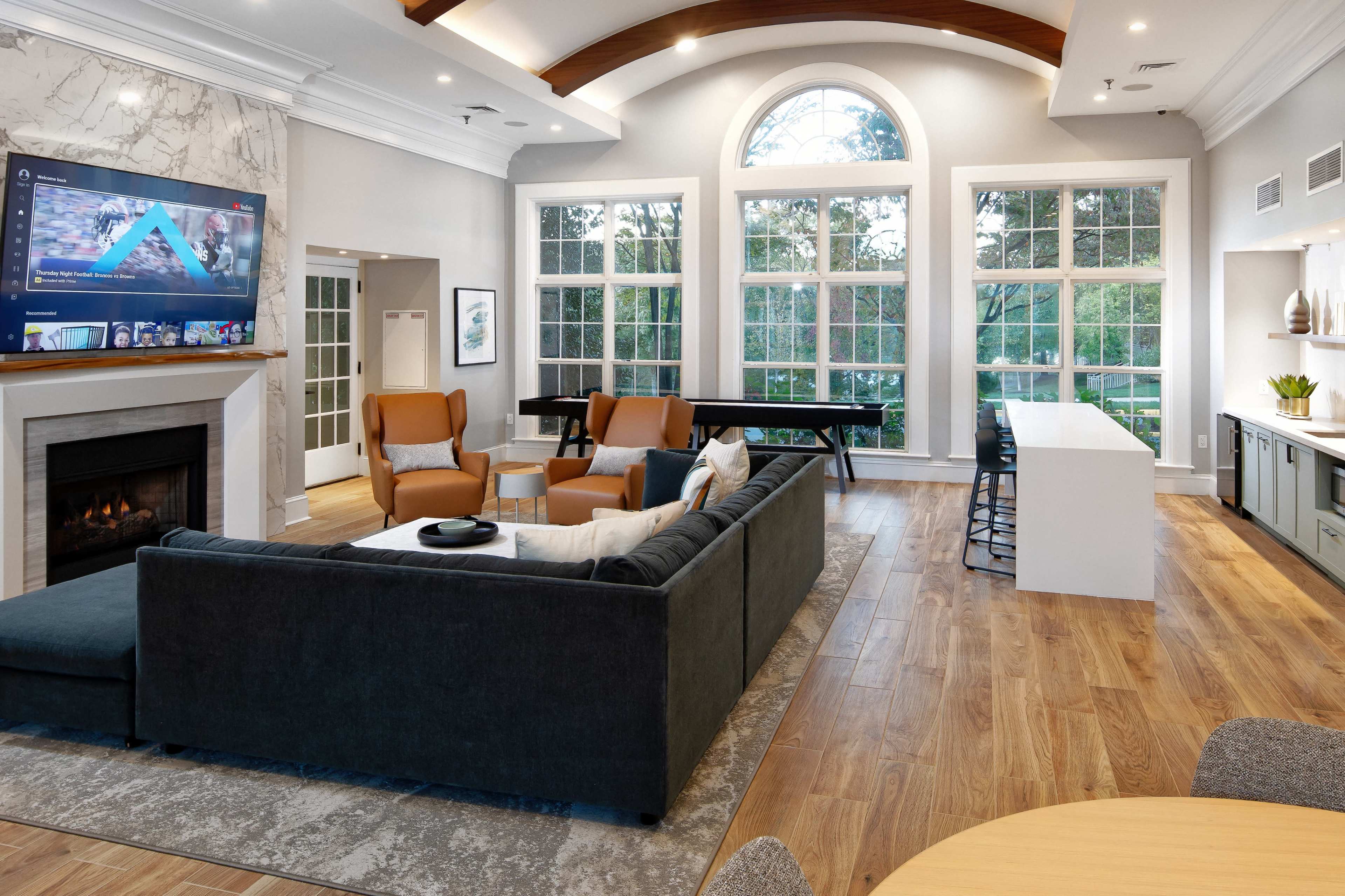 A spacious living room with a sectional sofa, a television above a fireplace, large windows, and a light-colored kitchen area with a dining table.