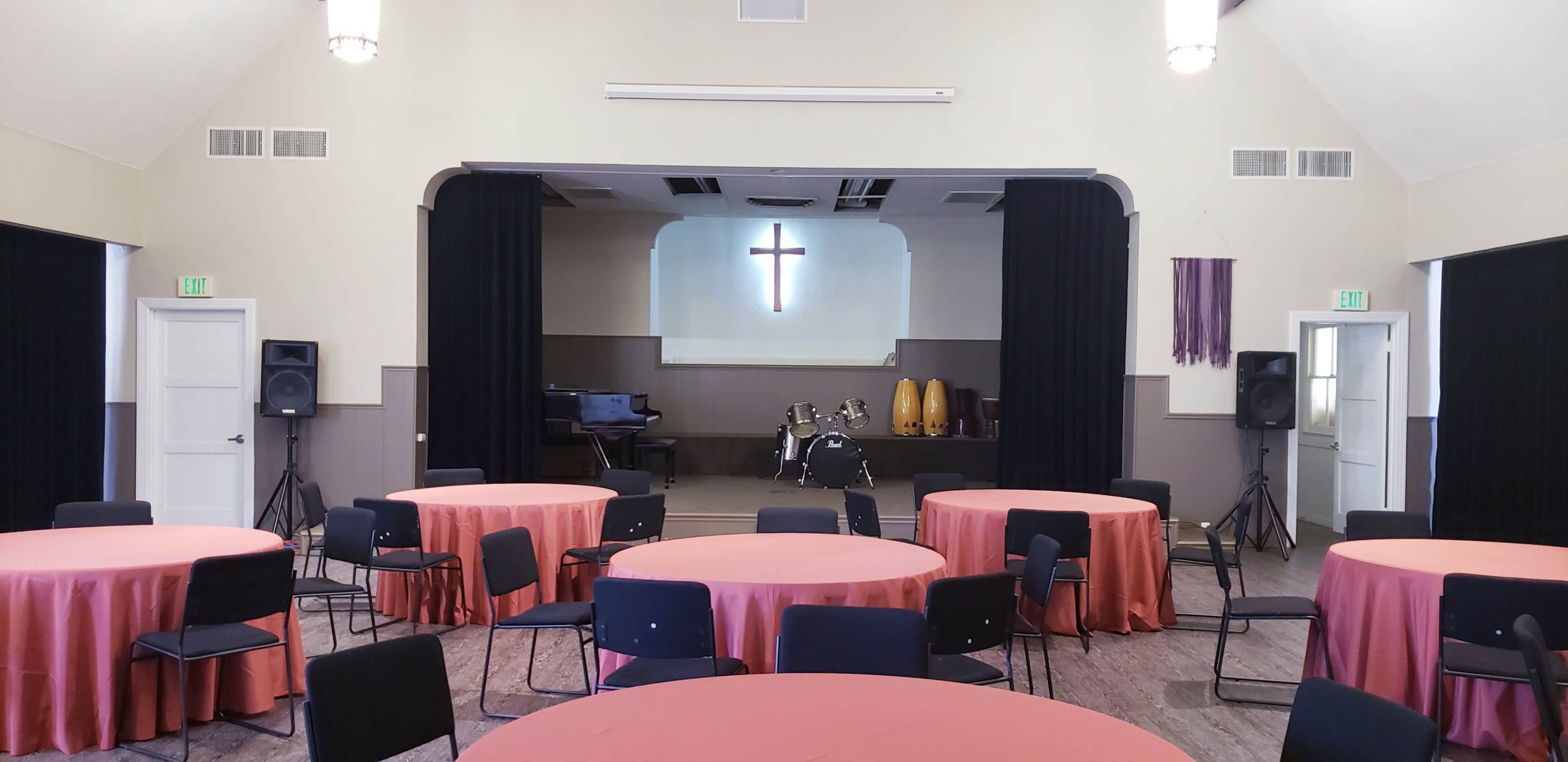 A hall set up for an event, featuring several round tables with orange tablecloths, chairs, and a stage with a cross backdrop and musical instruments.
