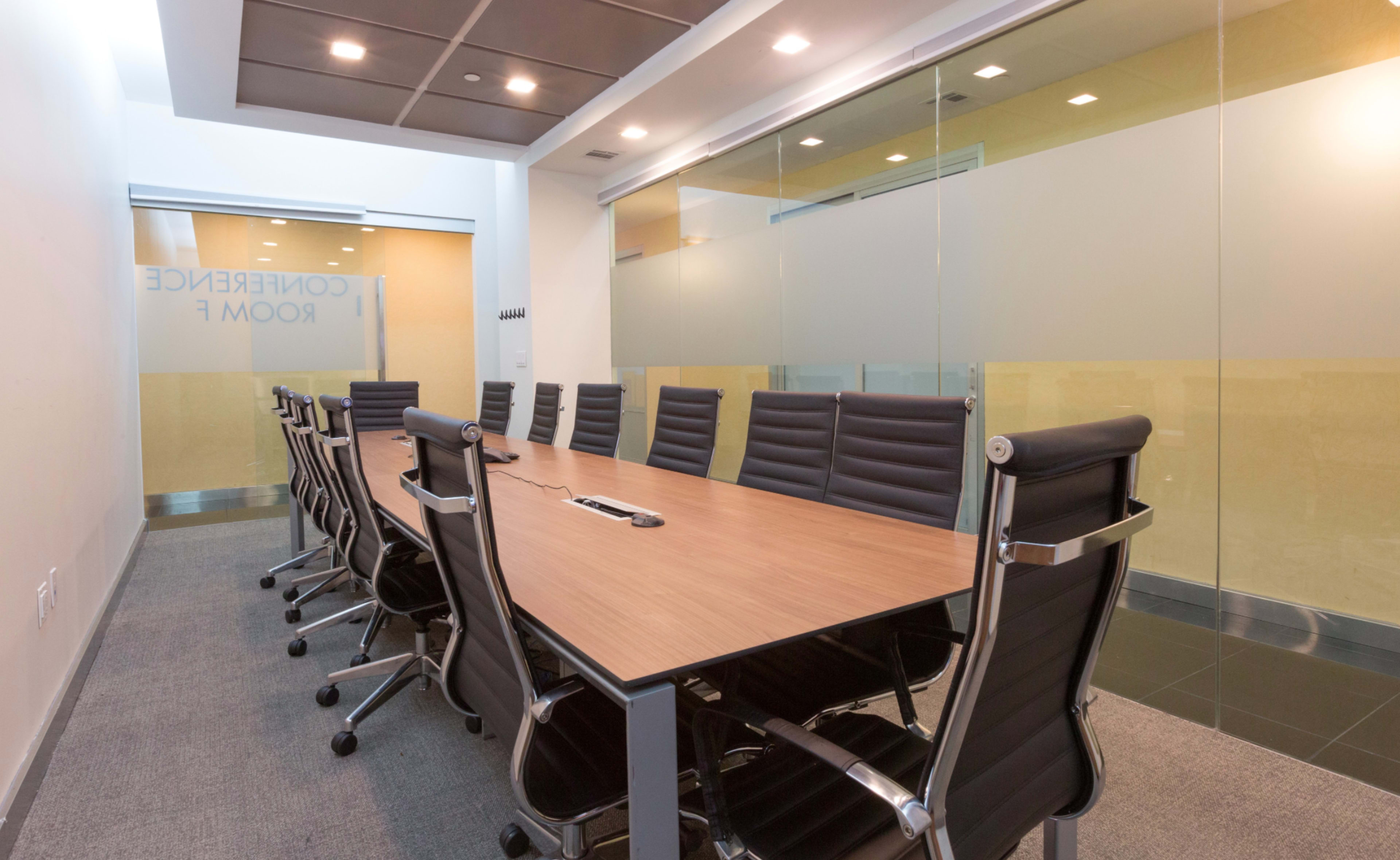 A spacious conference room features a long wooden table surrounded by black ergonomic chairs, with glass walls and modern lighting.