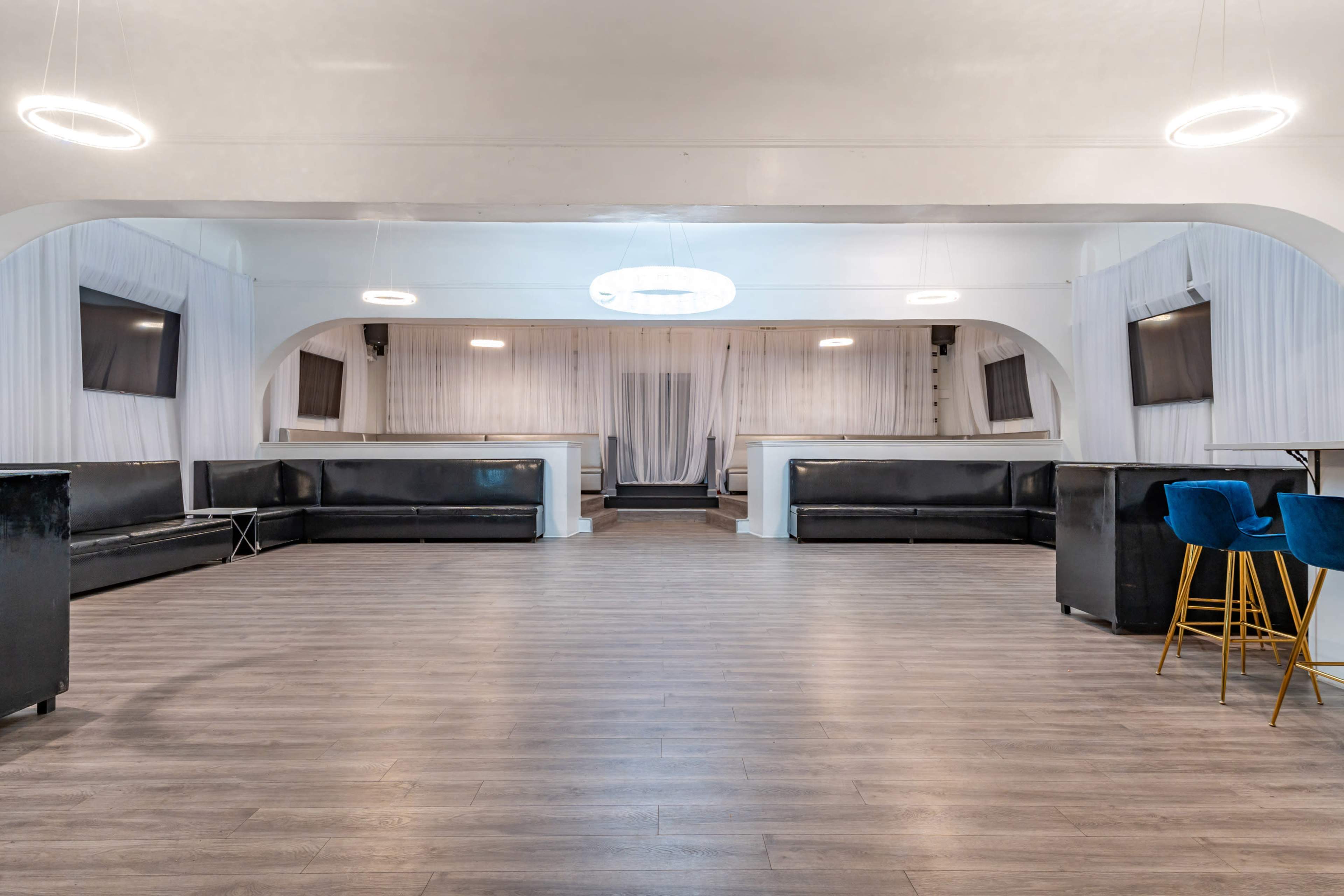 The image shows a spacious, modern event venue with a polished wooden floor, minimalist black seating, and white drapery in the background.