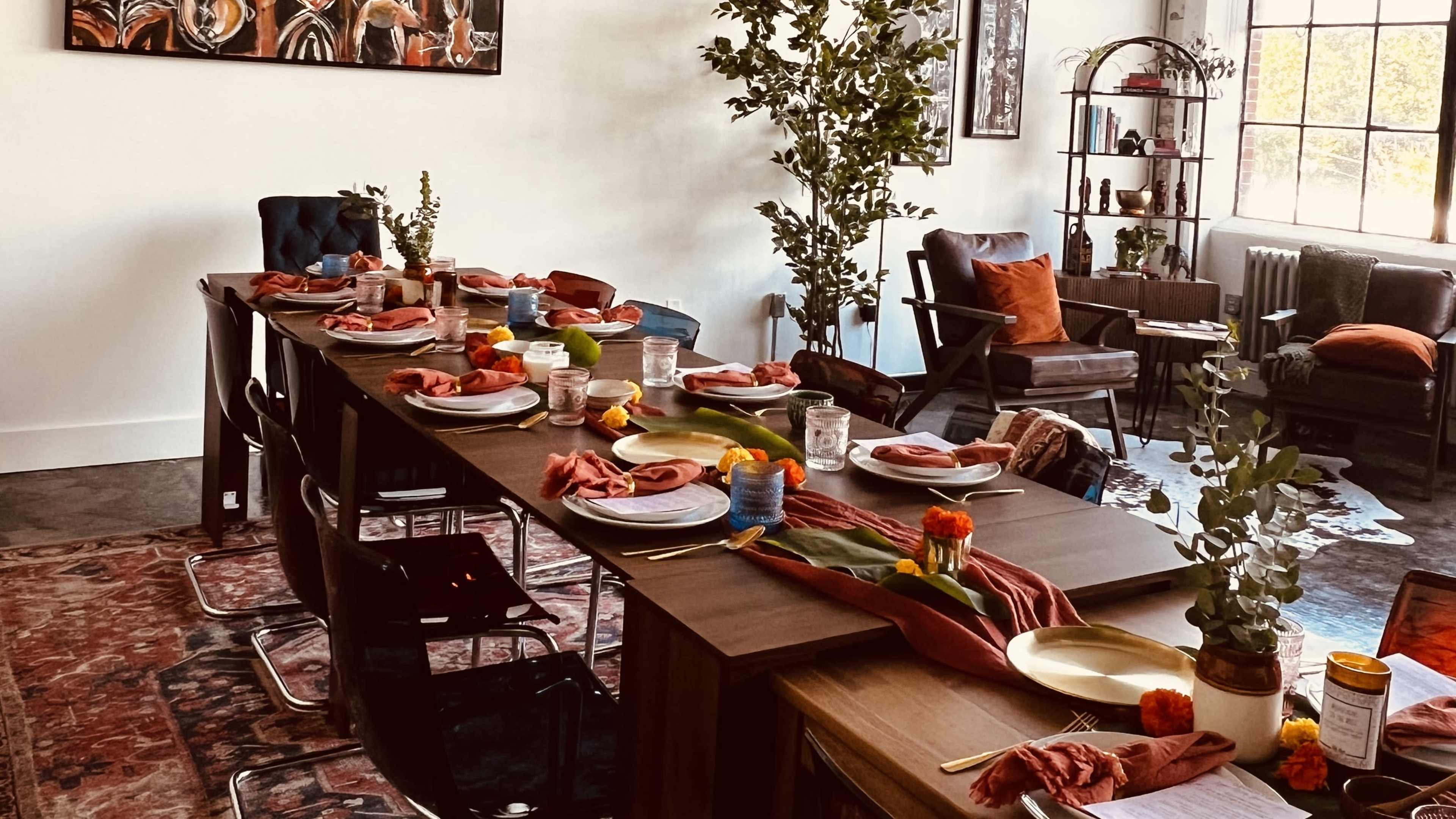 A long dining table is set with plates, napkins, and decorative flowers, surrounded by chairs in a well-lit room featuring a colorful rug and modern furnishings.