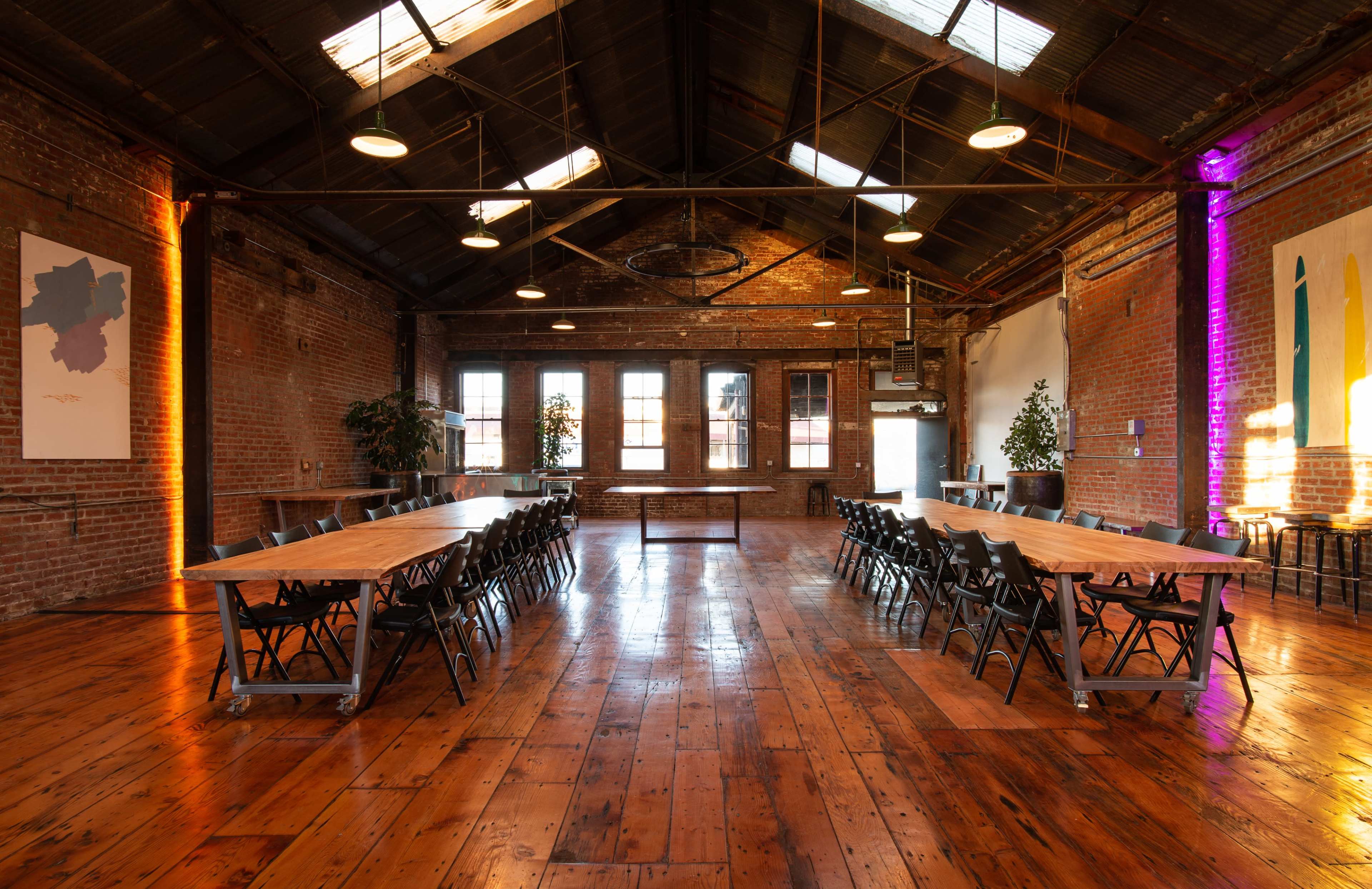 A spacious event room with high ceilings, exposed brick walls, and a long wooden table surrounded by black folding chairs.
