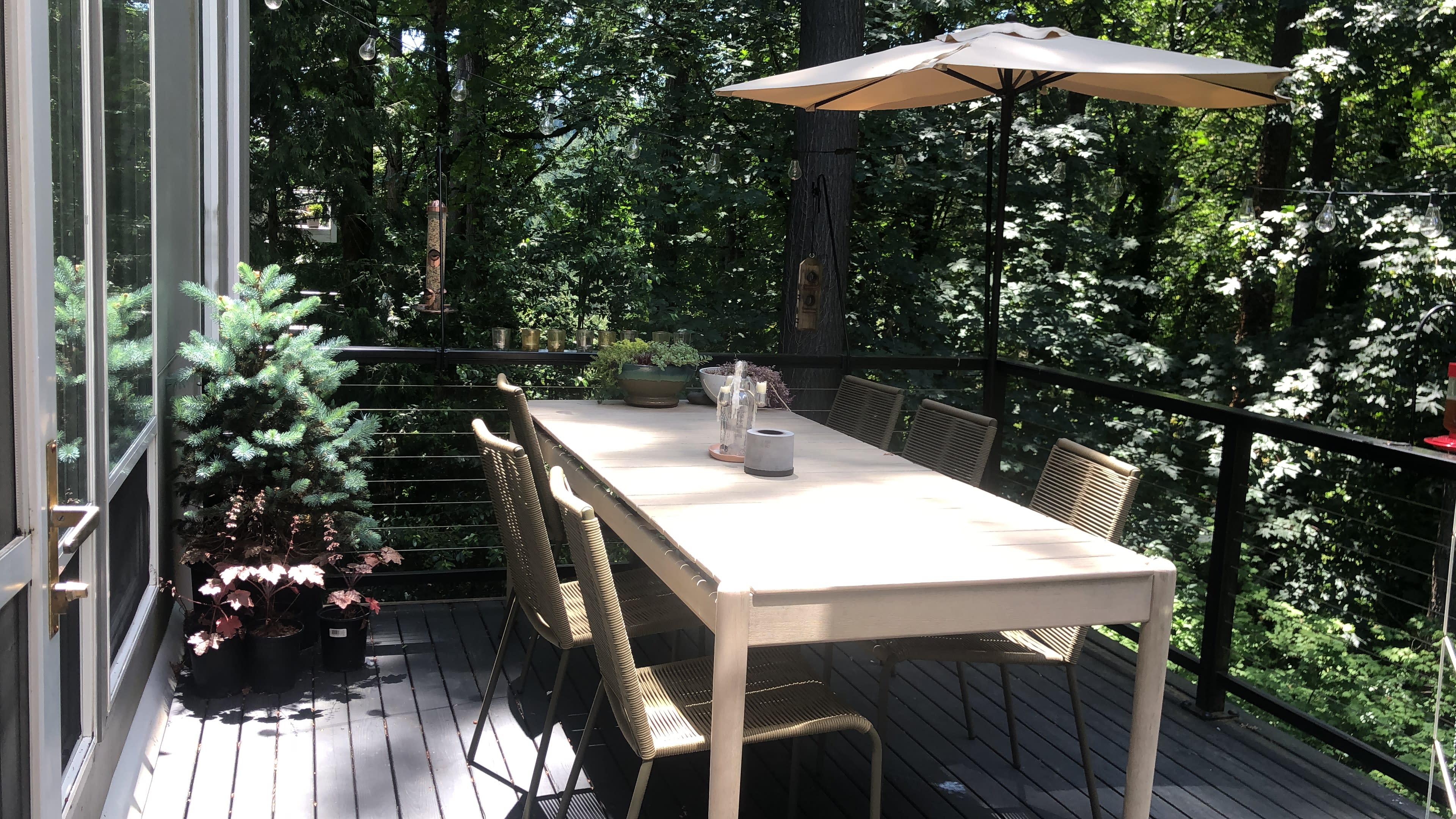 A long, white dining table with six chairs is set on a wooden deck surrounded by trees, featuring a small potted plant and an umbrella for shade.