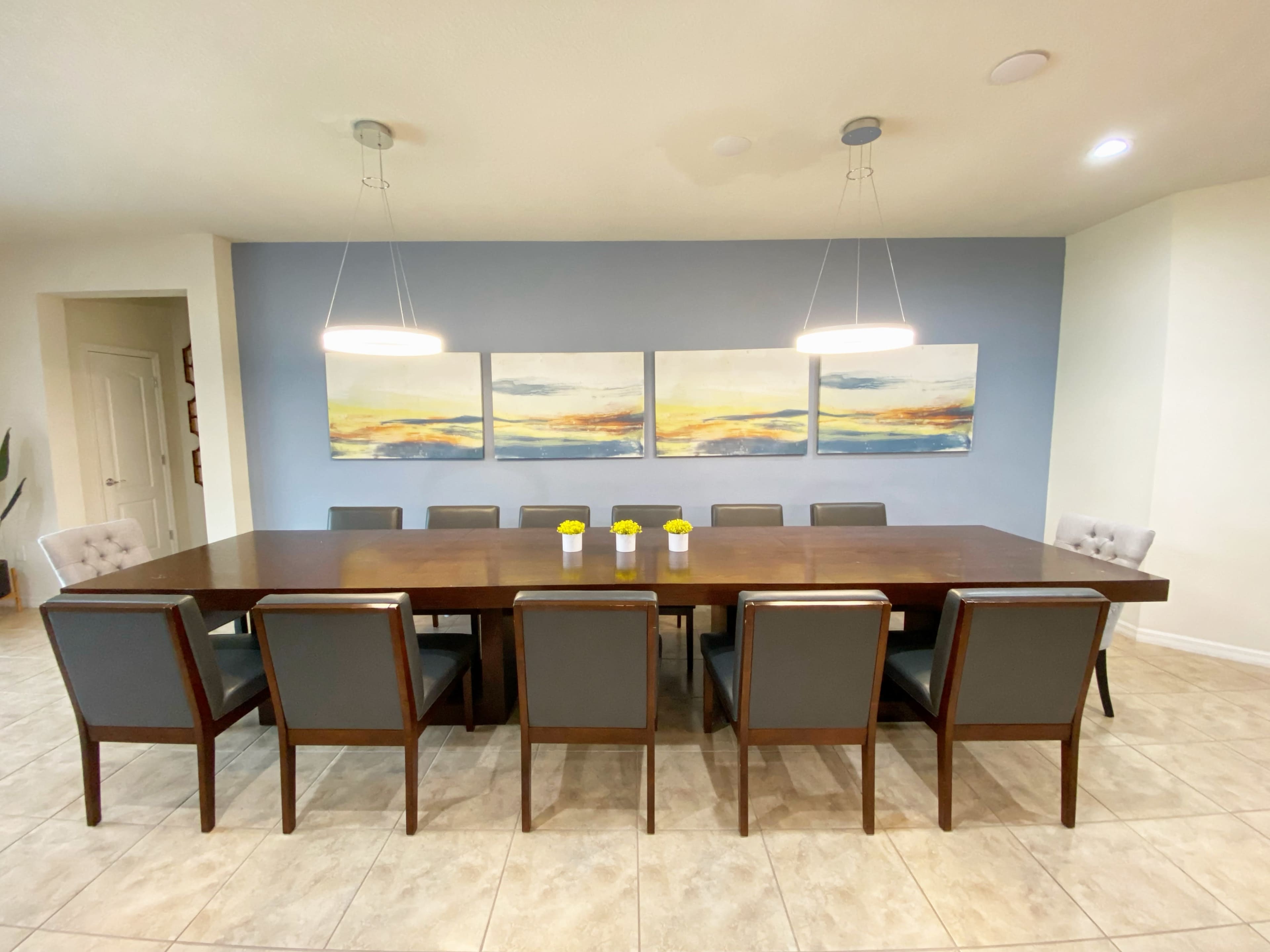 A spacious dining room features a long wooden table surrounded by ten chairs, with three paintings on the wall and two pendant lights above.