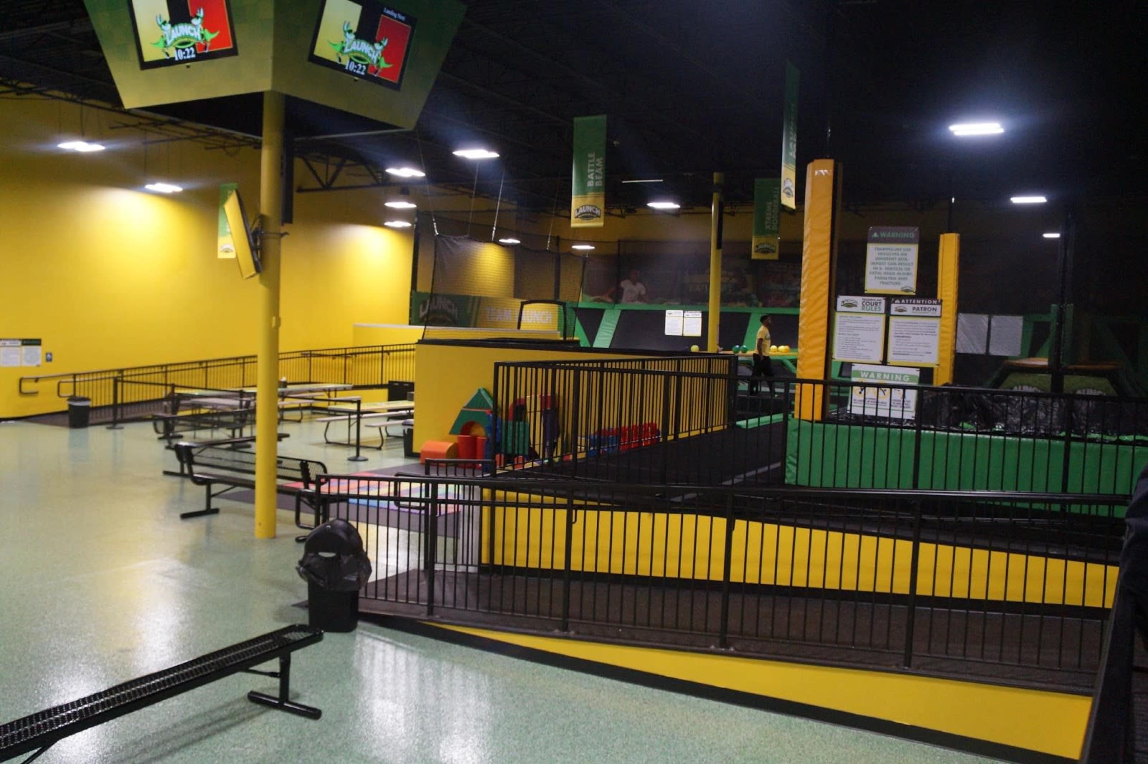 An indoor trampoline park with yellow walls, various play areas, seating benches, and multiple safety nets.