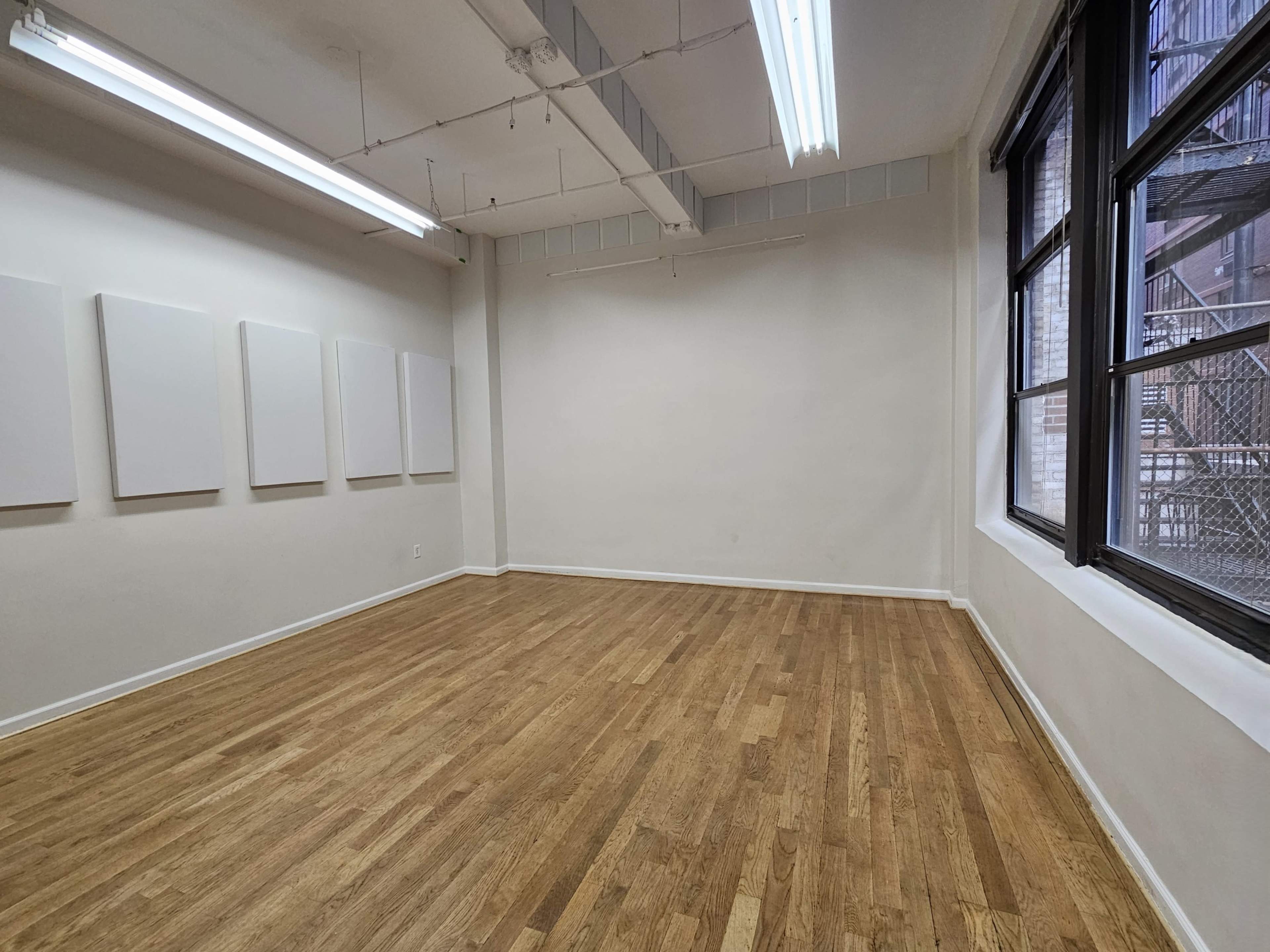 The image shows a vacant art studio with wooden flooring and blank white walls, featuring large windows that overlook an external metal fire escape.