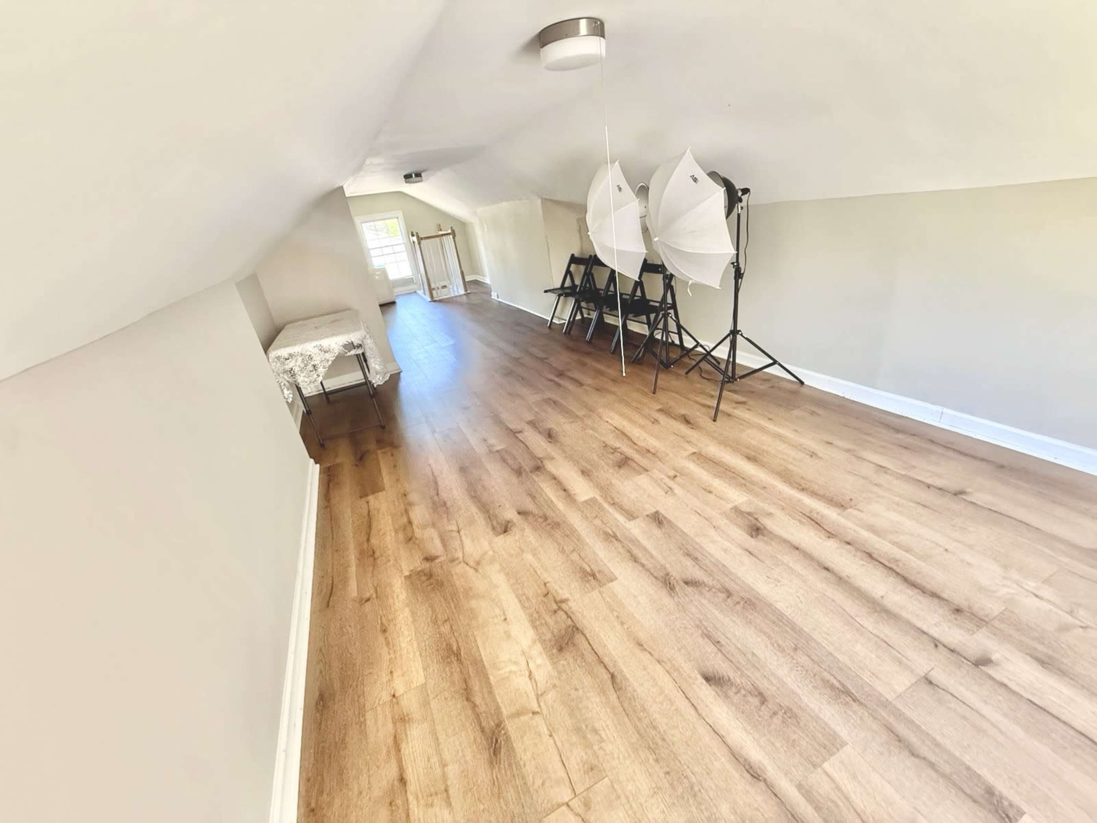 The image shows a narrow, well-lit attic space with wooden flooring, two folding chairs, and photography lighting equipment set up at one end.