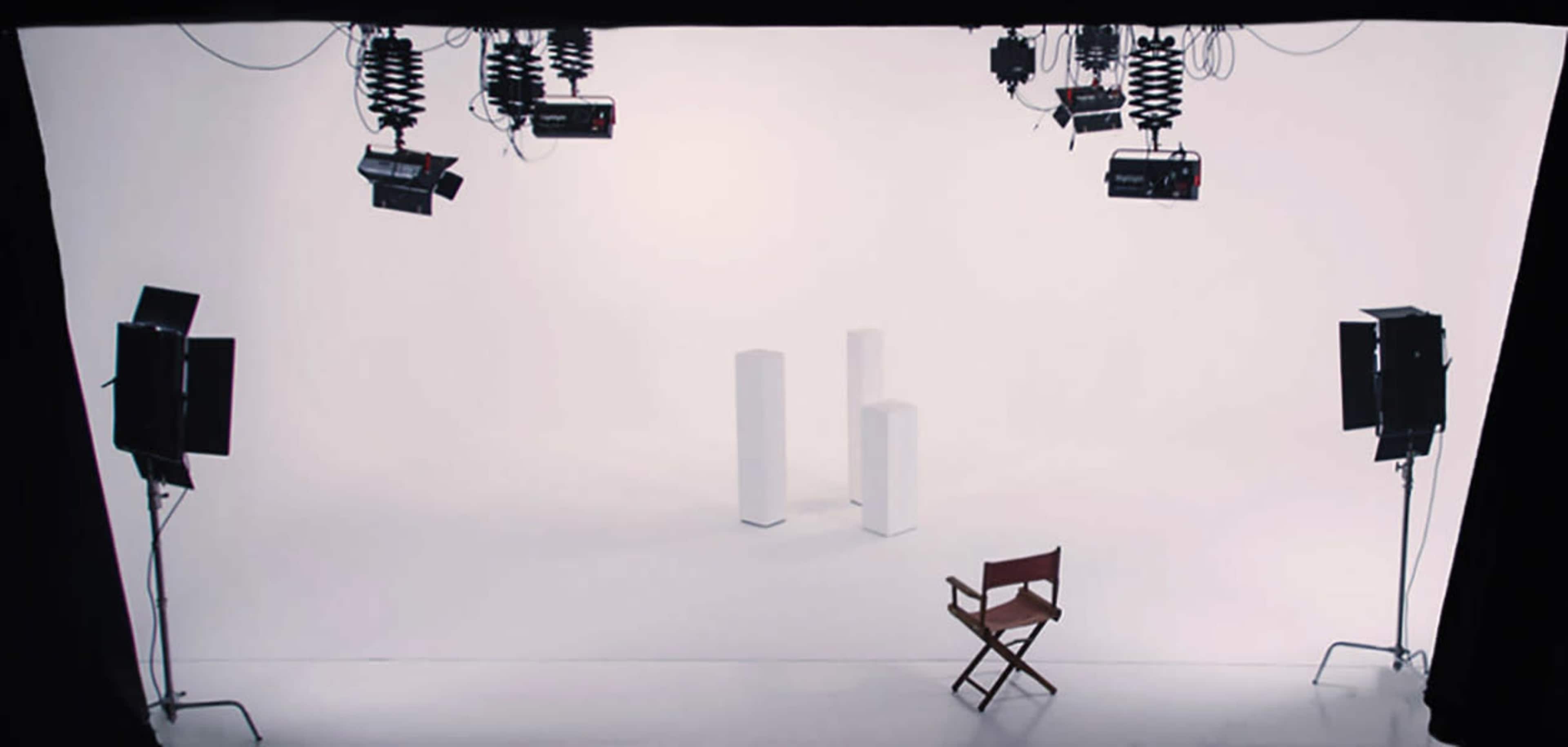A white studio backdrop with two light stands, two light sources, and a wooden director's chair placed in front of two white pedestals.