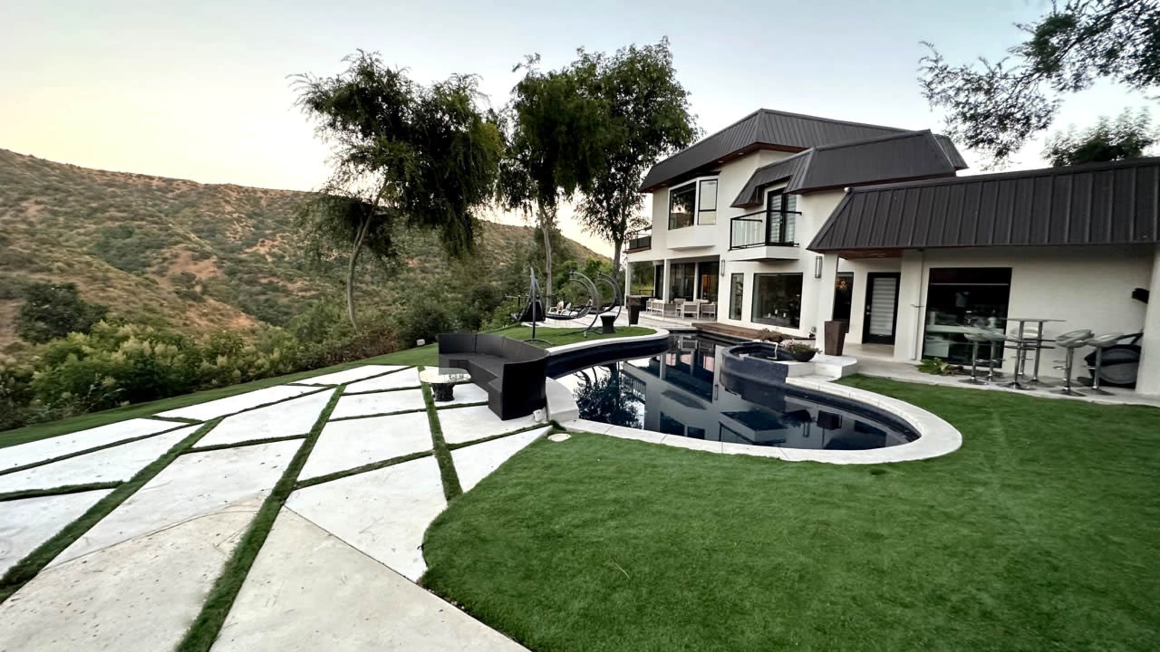 A modern house with large windows overlooks a swimming pool and landscaped yard, set against a hilly backdrop.