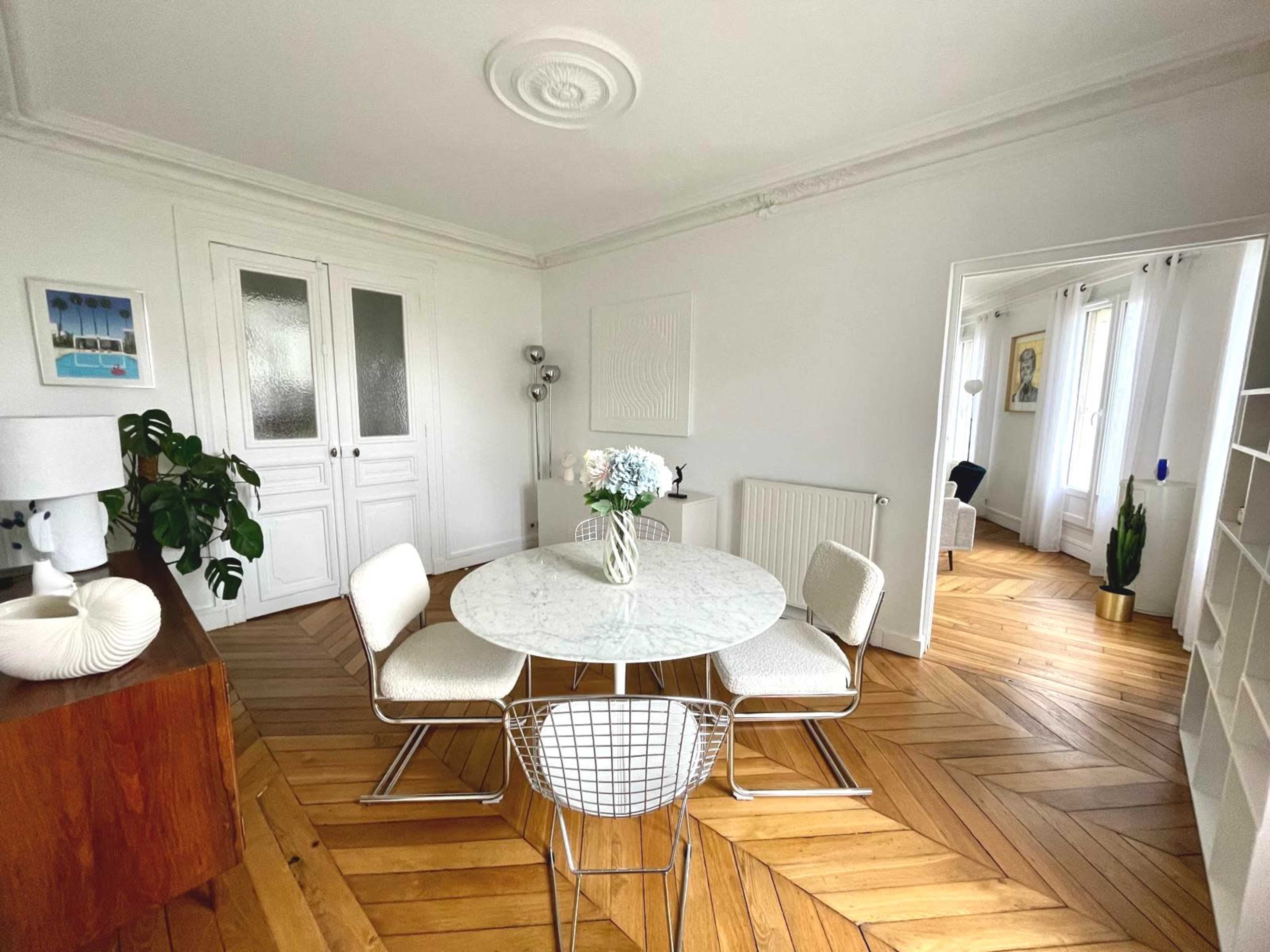 The image shows a modern dining area with a round marble table and four chairs on a hardwood floor, featuring white walls and natural light from nearby windows.