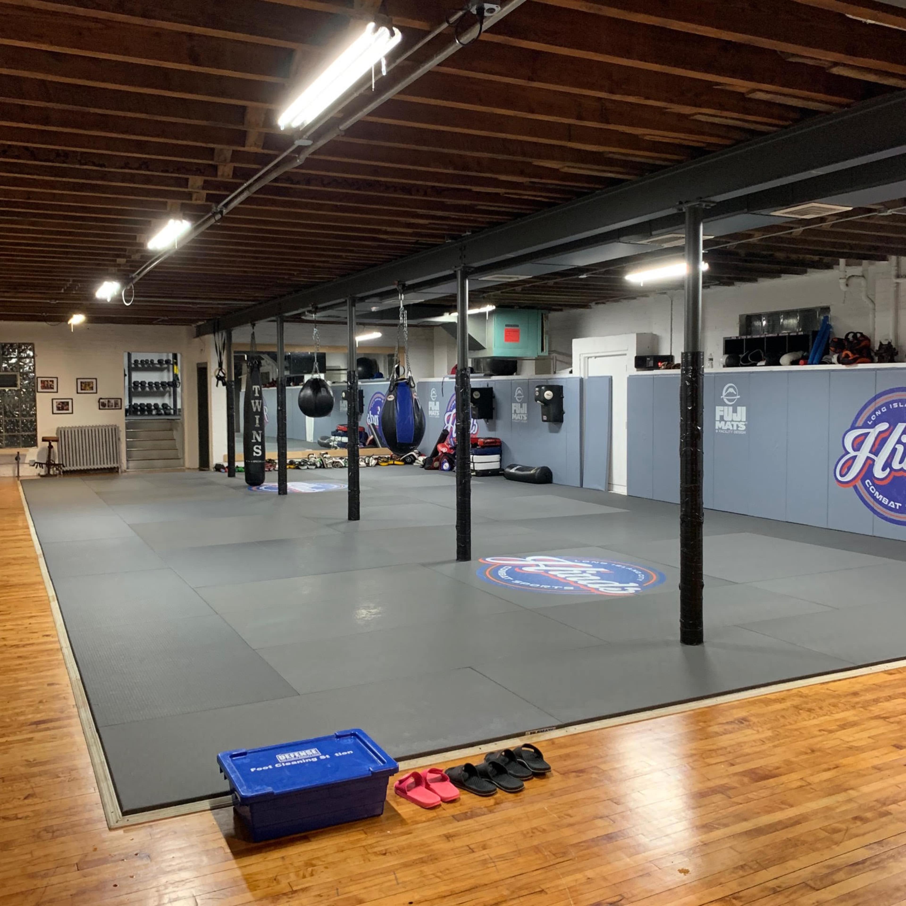 A spacious martial arts training area with padded flooring, hanging punching bags, and gym equipment along the walls.