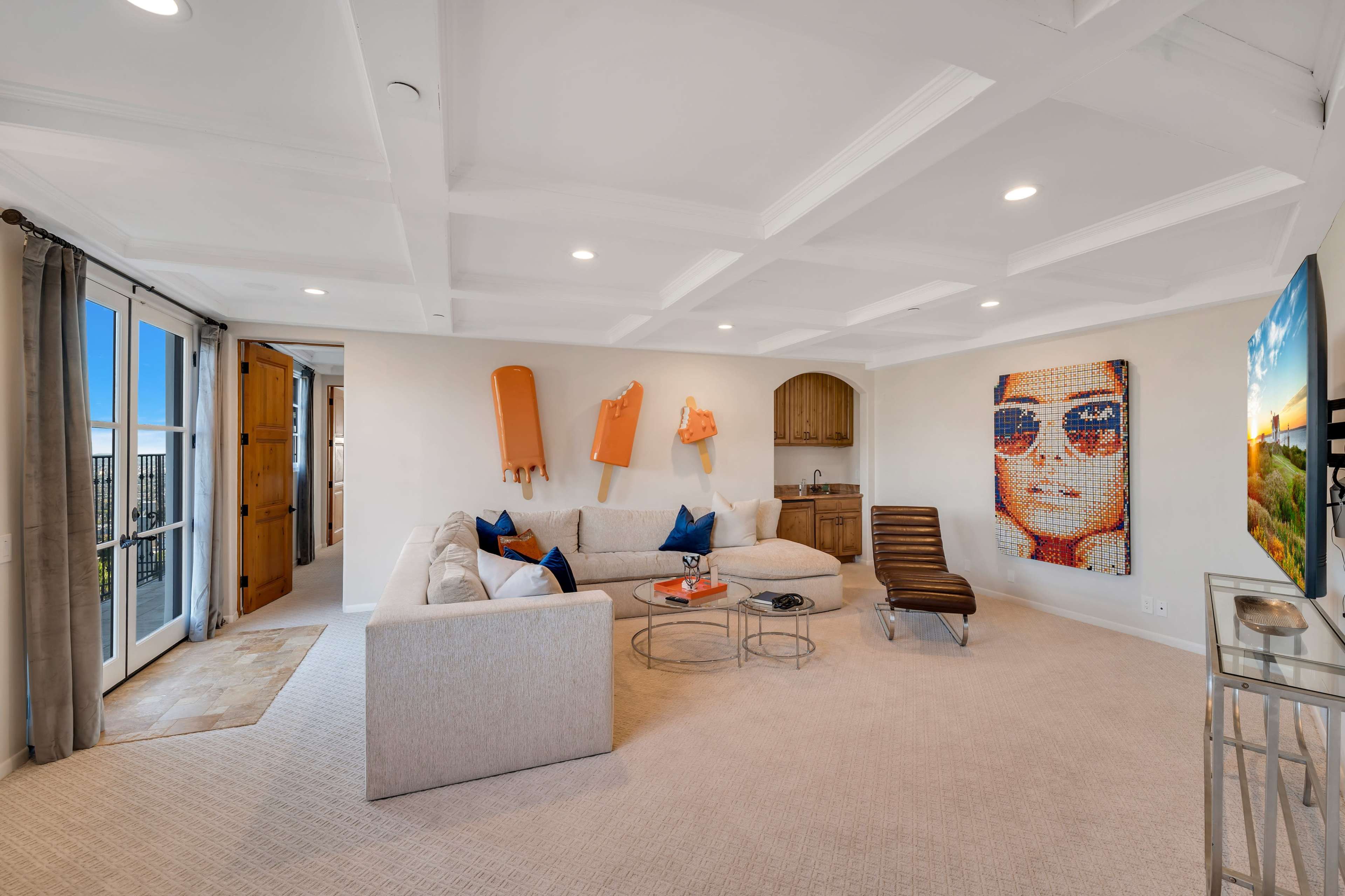 The room features a spacious living area with a large sectional sofa, a modern chair, and a wall art piece of a person wearing sunglasses.