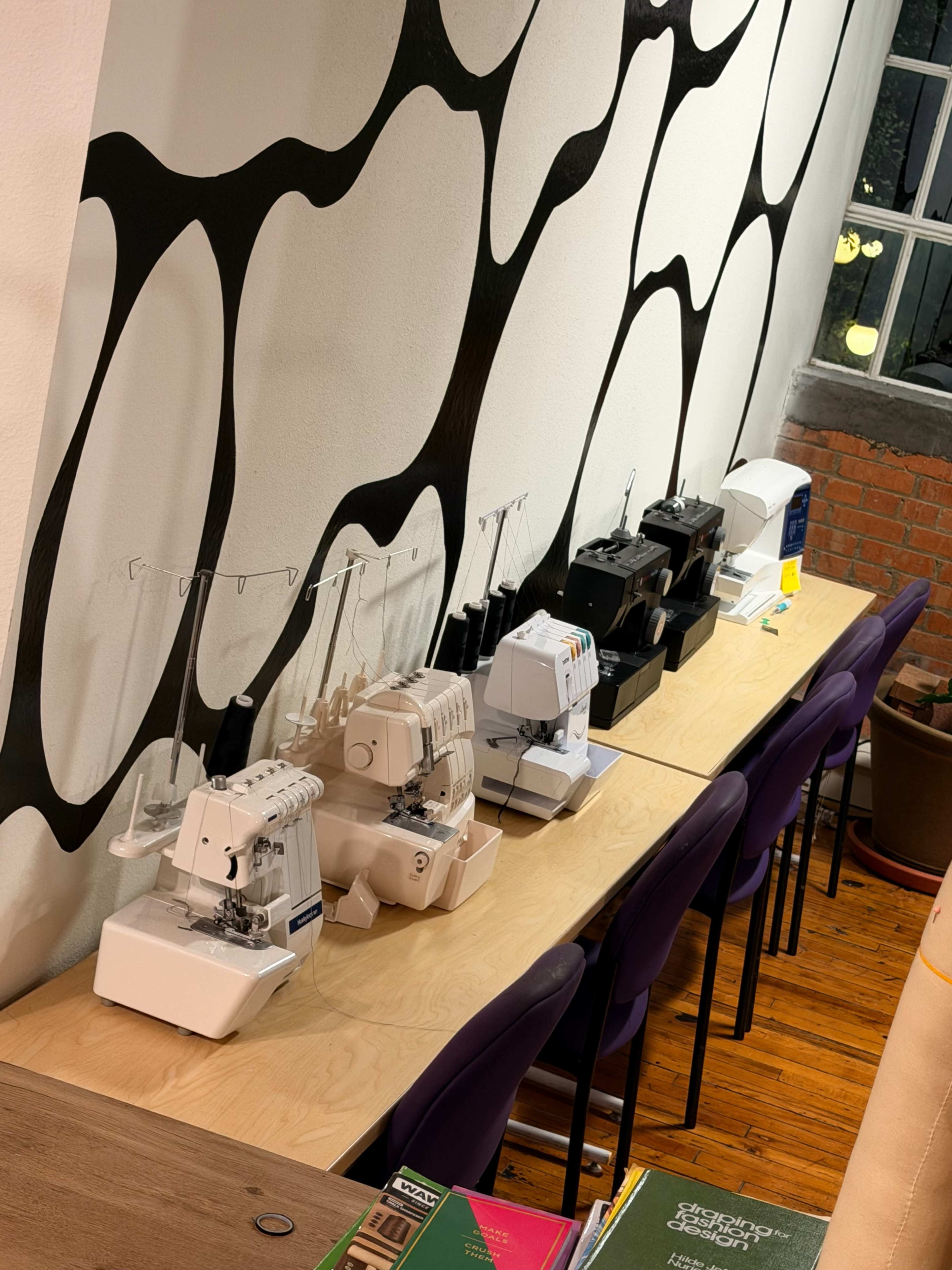 The image shows a row of sewing machines lined up on a wooden table against a patterned wall.