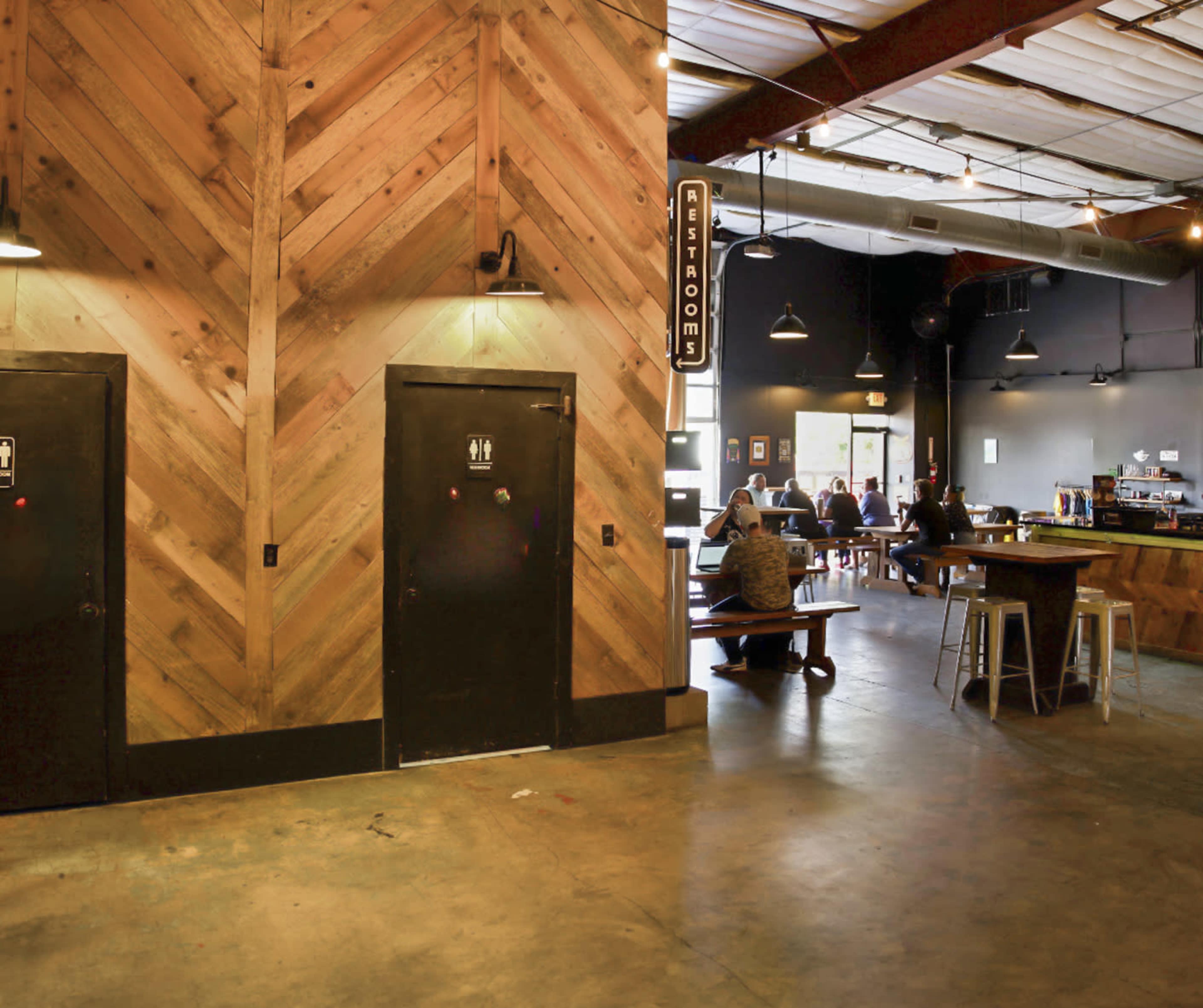 A spacious dining area with wooden accents, featuring two restroom doors on the left and several tables with patrons seated at the back.
