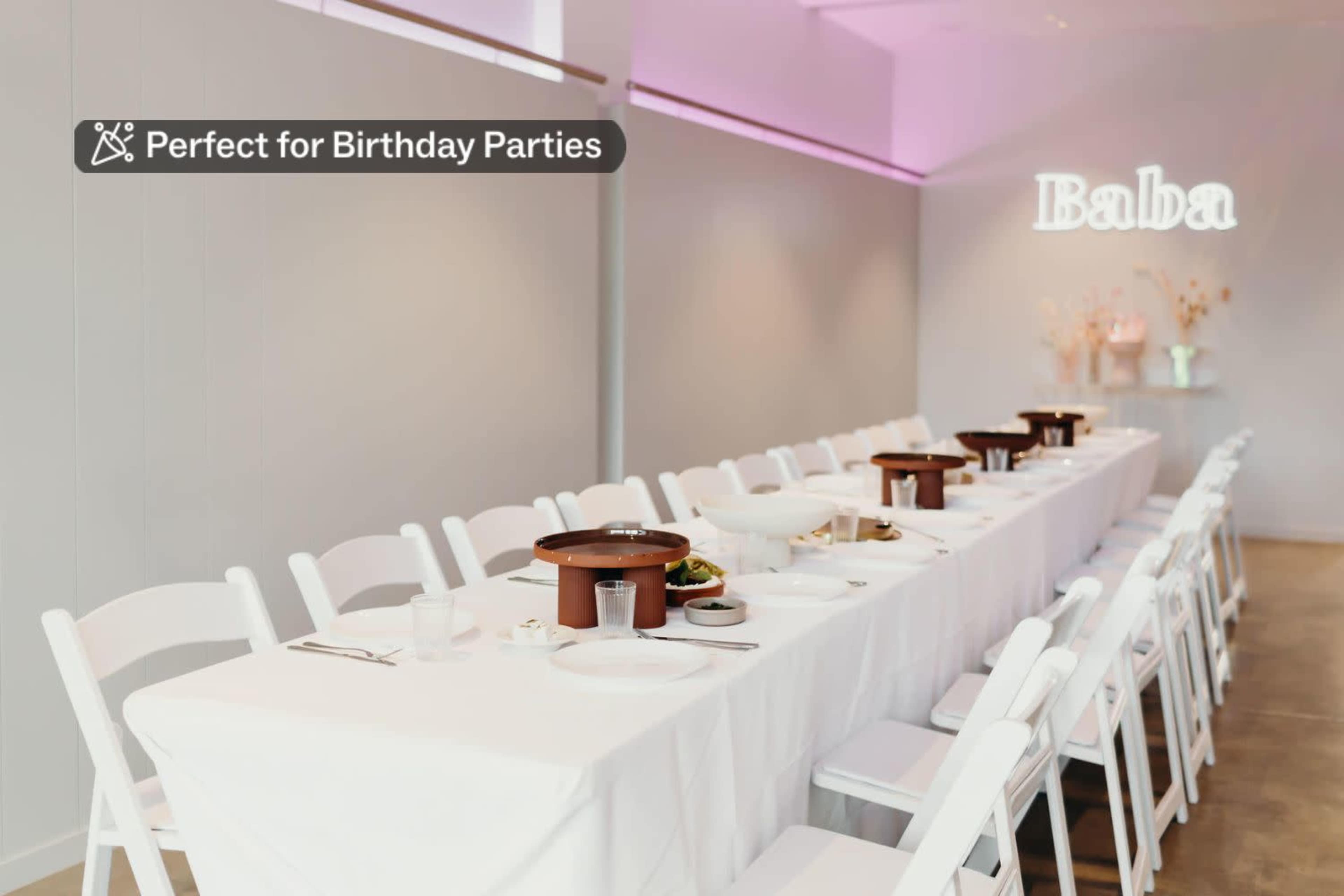 A long, white table is set for a birthday party with plates and bowls, and a neon sign that reads "Baba" on the wall.